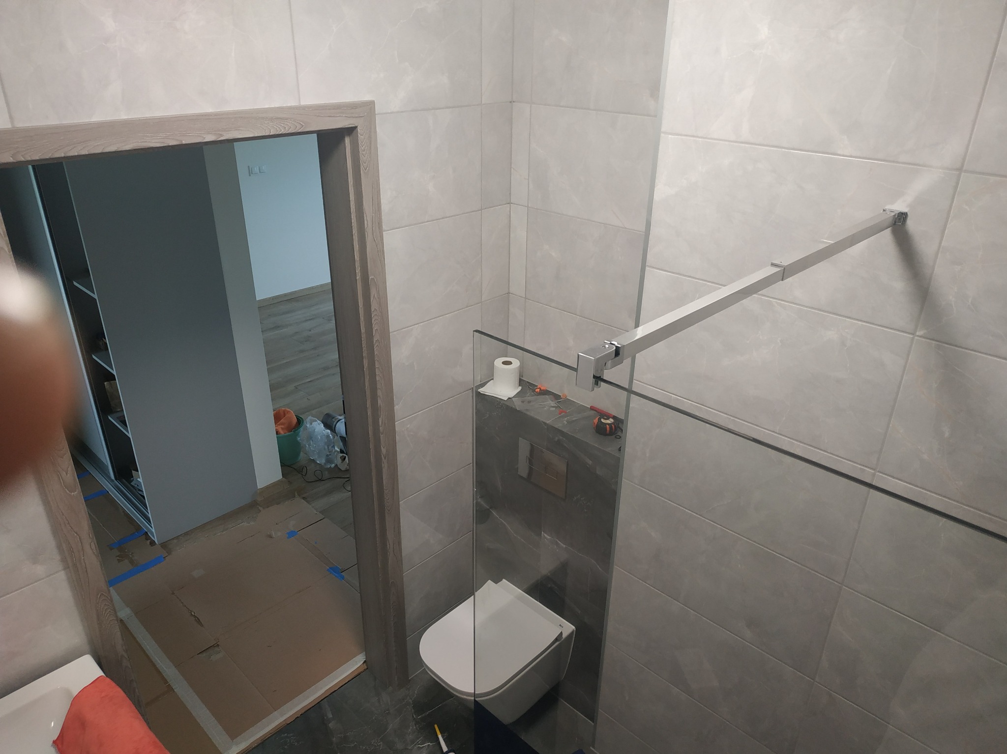 Wykończona łazienka z szarymi płytkami, kabiną prysznicową ze szklaną ścianką i podwieszaną toaletą. Widoczne narzędzia i akcesoria, sugerujące świeży remont.