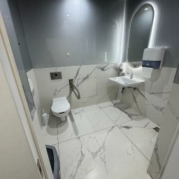 Piotr STARK - Błyszcząca, czysta toaleta z marmurowymi płytkami, podświetlanym lustrem i nowoczesną armaturą. Widok zza uchylonych drzwi.