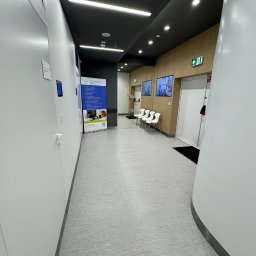 Piotr STARK - Wnętrze nowoczesnego korytarza biurowego z jasną podłogą, białymi ścianami i ciemnym sufitem. Widoczne plakaty informacyjne i krzesła dla oczekujących gości.
