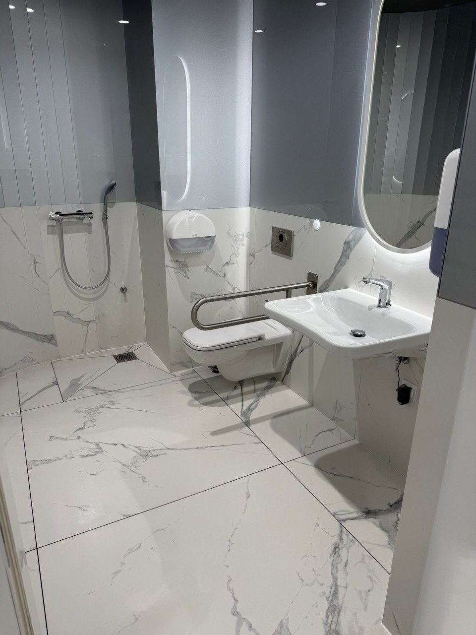 Biała, luksusowa łazienka z marmurową podłogą i ścianami. Umywalka, toaleta dla niepełnosprawnych, lustro z podświetleniem. Czystość i minimalizm.