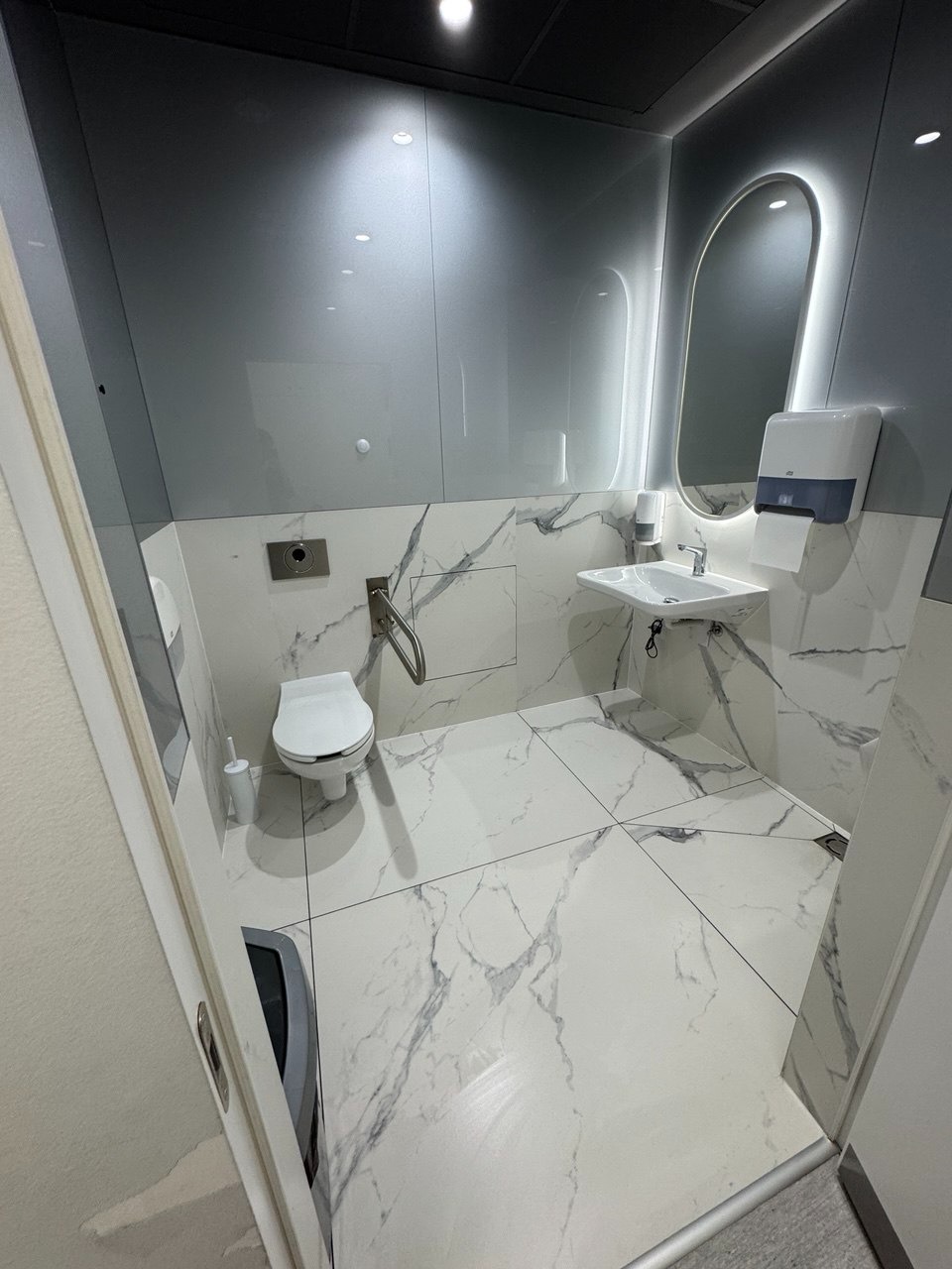 Błyszcząca, czysta toaleta z marmurowymi płytkami, podświetlanym lustrem i nowoczesną armaturą. Widok zza uchylonych drzwi.