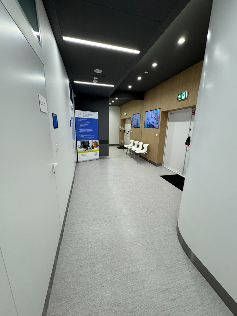 Wnętrze nowoczesnego korytarza biurowego z jasną podłogą, białymi ścianami i ciemnym sufitem. Widoczne plakaty informacyjne i krzesła dla oczekujących gości.