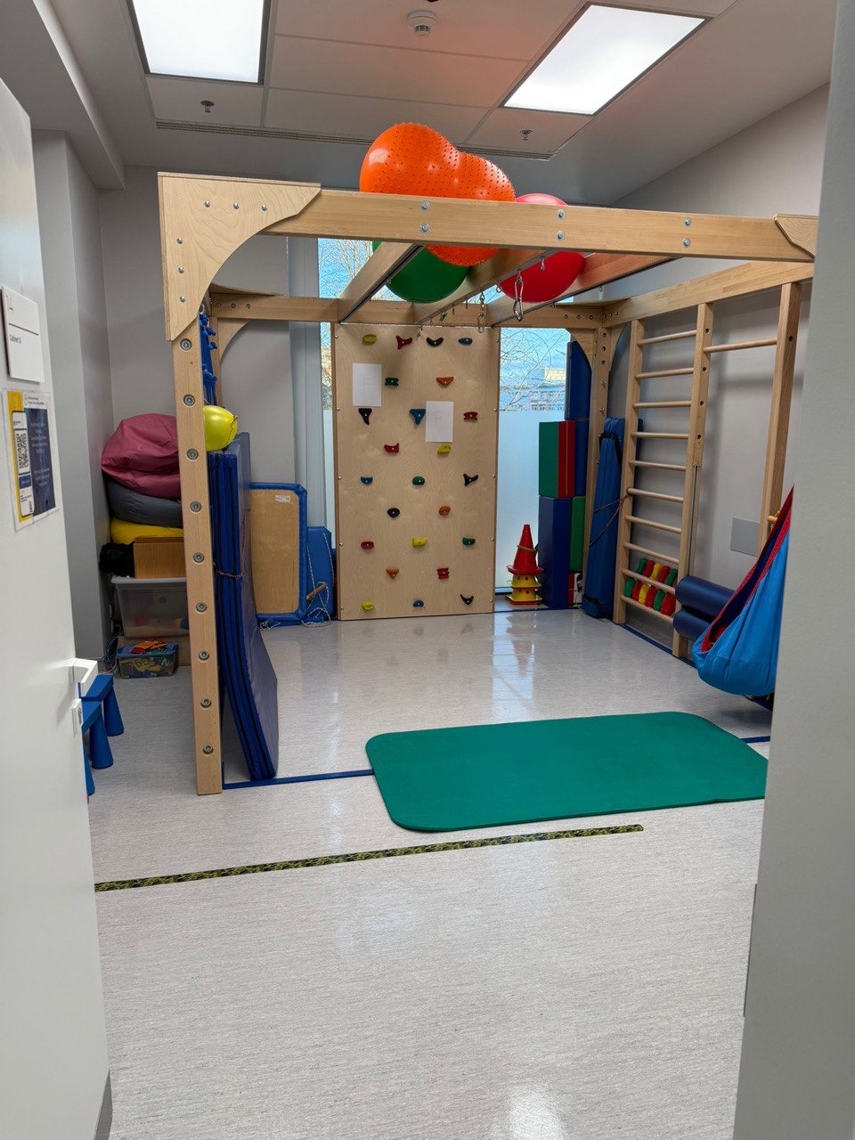 Sala sensoryczna z drabinkami, ścianką wspinaczkową i kolorowymi piłkami. Widok zza progu, czysta podłoga, jasne oświetlenie.