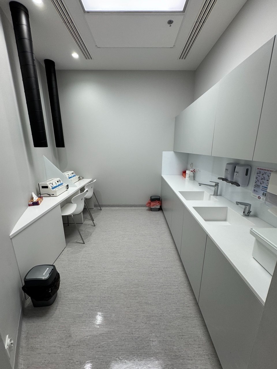 Jasne wnętrze gabinetu medycznego z umywalkami, białymi szafkami i sprzętem. Dwa czarne, pionowe przewody wentylacyjne przy ścianie. Czysta podłoga i minimalistyczny design.