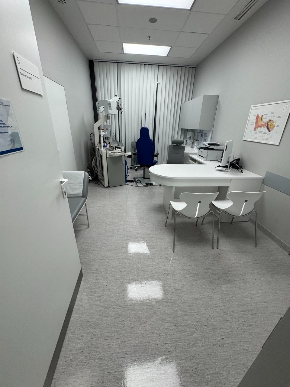 Wnętrze gabinetu lekarskiego z białymi meblami, specjalistycznym sprzętem medycznym i lśniącą podłogą. Widoczne krzesła, biurko i schemat ucha na ścianie.