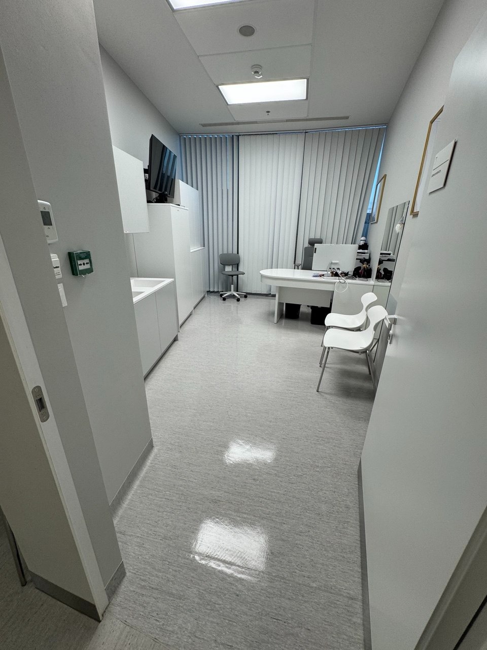 Wnętrze biura z białymi meblami, biurkiem i krzesłami, widok z perspektywy wejścia. Jasne, neutralne kolory, minimalistyczny design, czyste podłogi z odbiciami światła.