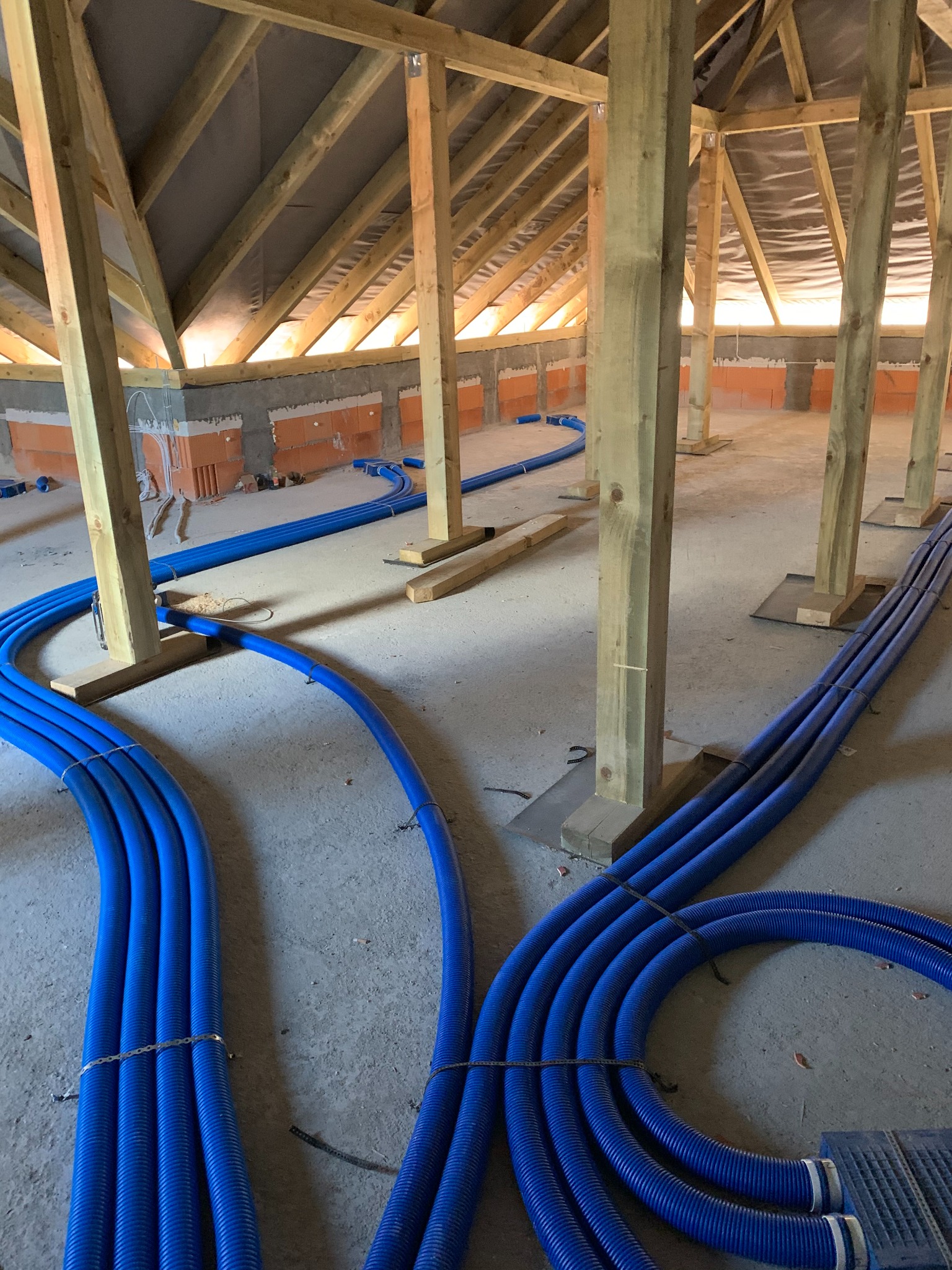 Niebieskie rury ogrzewania podłogowego ułożone w surowym wnętrzu poddasza z drewnianą konstrukcją. Widoczne ceglane ściany i izolacja dachu.