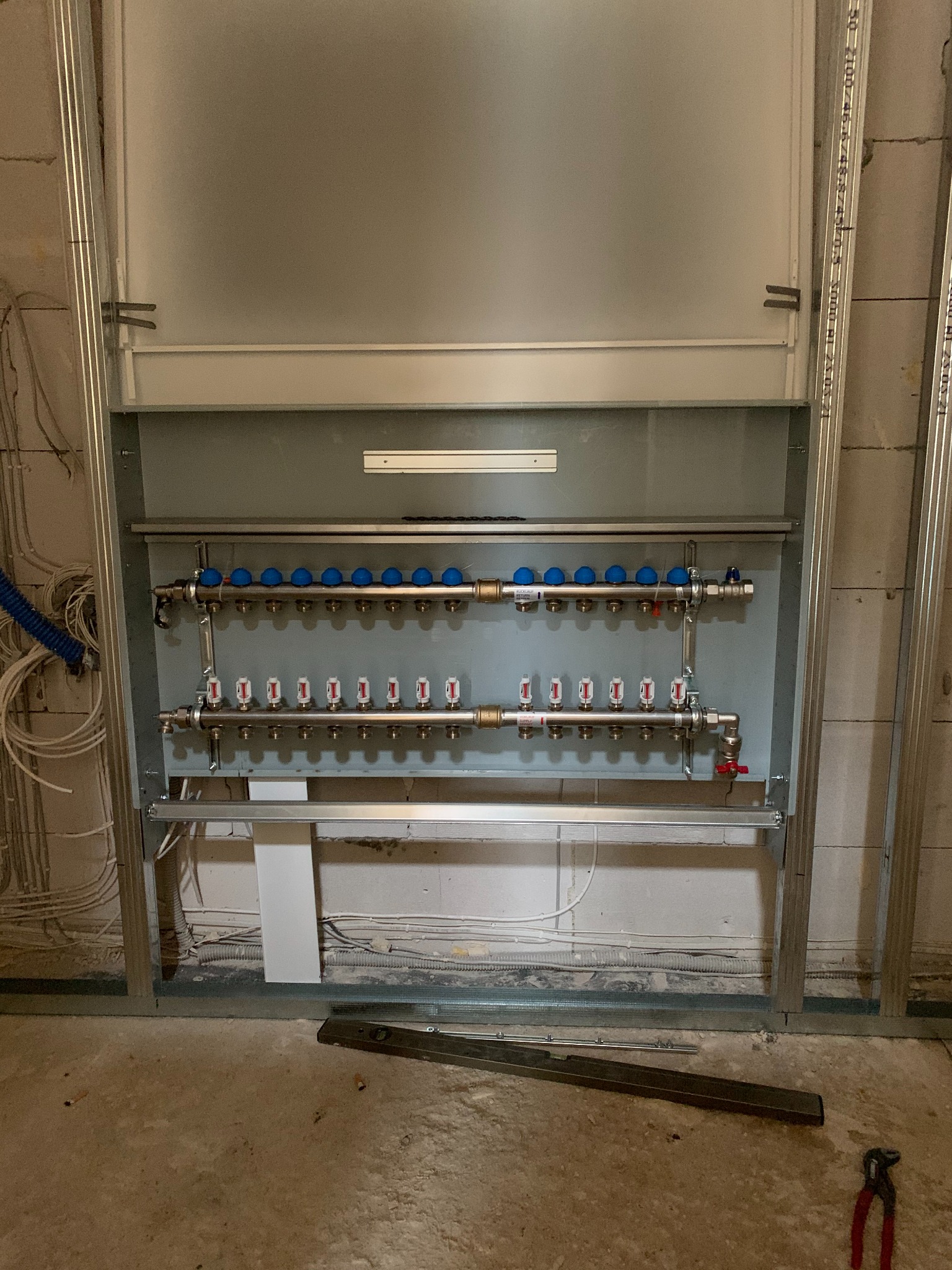 Rozdzielacz ogrzewania podłogowego w metalowej szafce, widoczne rury, zawory i liczniki. Surowy stan wykończenia ścian i podłogi. Narzędzia na podłodze.