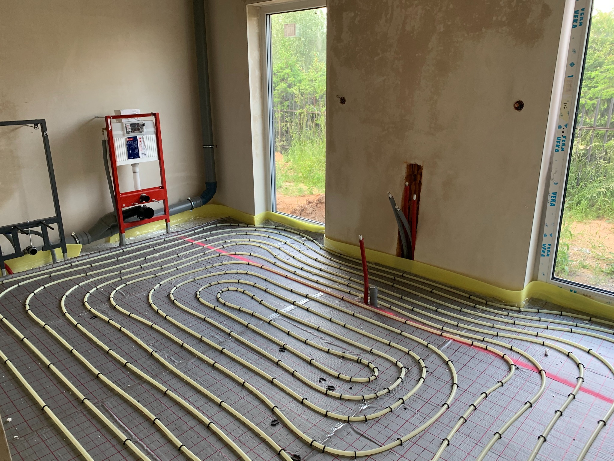 Ułożone rury ogrzewania podłogowego na folii izolacyjnej w surowym wnętrzu z oknem. Widoczny stelaż podtynkowy WC i pion kanalizacyjny.