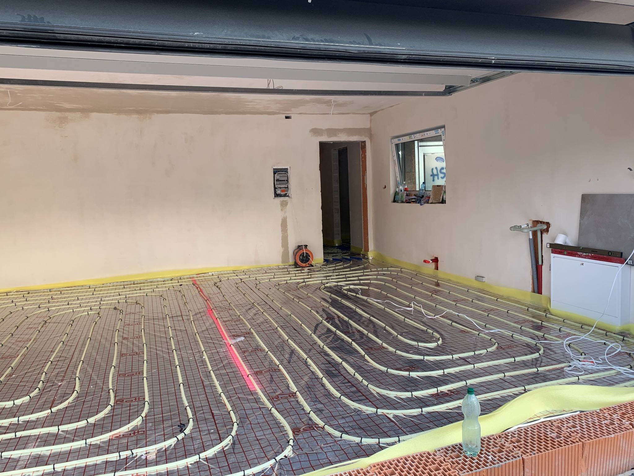 Ułożona instalacja ogrzewania podłogowego na żółtej macie izolacyjnej w surowym wnętrzu. Widoczne rury grzewcze, siatka montażowa i butelka wody.