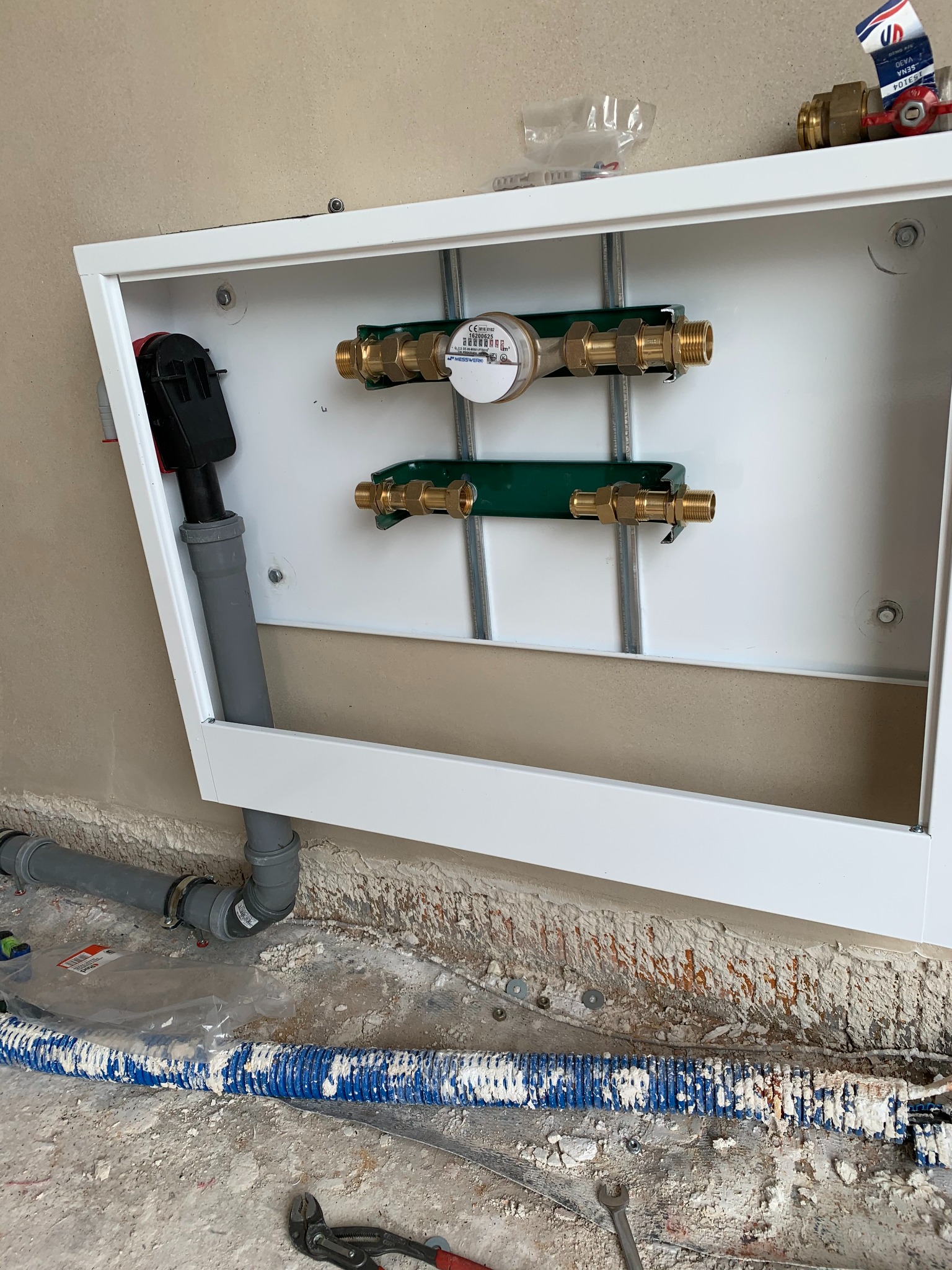 Zamknięta biała skrzynka z zamontowanym wodomierzem i rurami z mosiężnymi złączkami. Na podłodze widoczne rury i narzędzia hydrauliczne.
