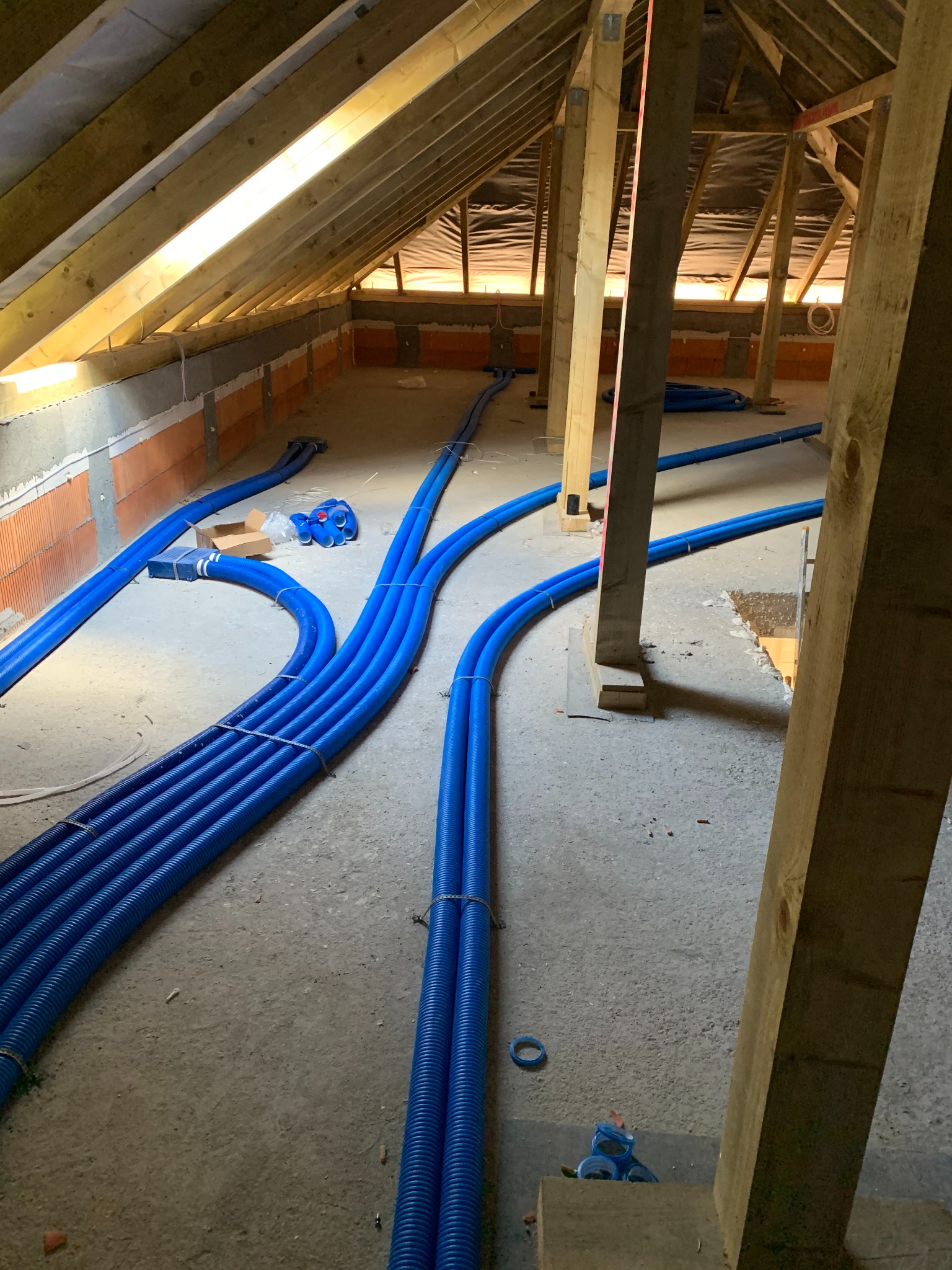 Niebieskie rury instalacji podłogowej ułożone w surowym wnętrzu poddasza z drewnianą konstrukcją dachu. Widoczne elementy mocujące i fragmenty ścian z cegły.