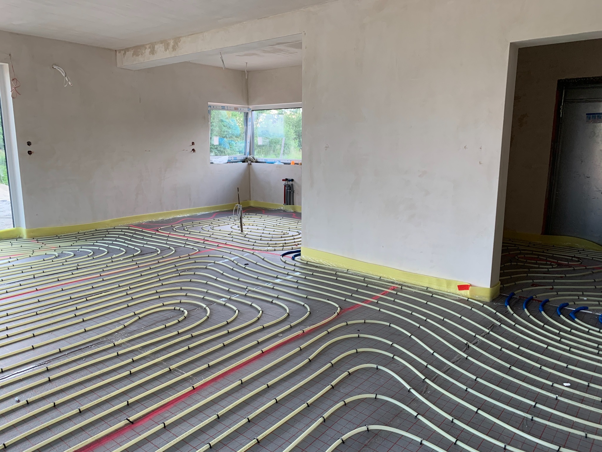Ułożone rury ogrzewania podłogowego na siatce w surowym wnętrzu. Widoczne żółte taśmy przy ścianach i fragment okna z zielenią na zewnątrz.