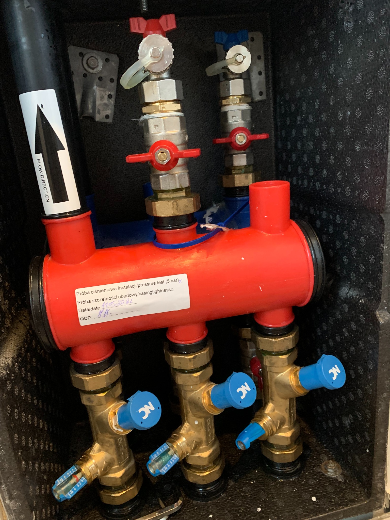 Czerwony rozdzielacz instalacji z zaworami oznaczonymi kolorami, umieszczony w czarnej obudowie. Widoczna etykieta z datą próby ciśnieniowej i szczelności.