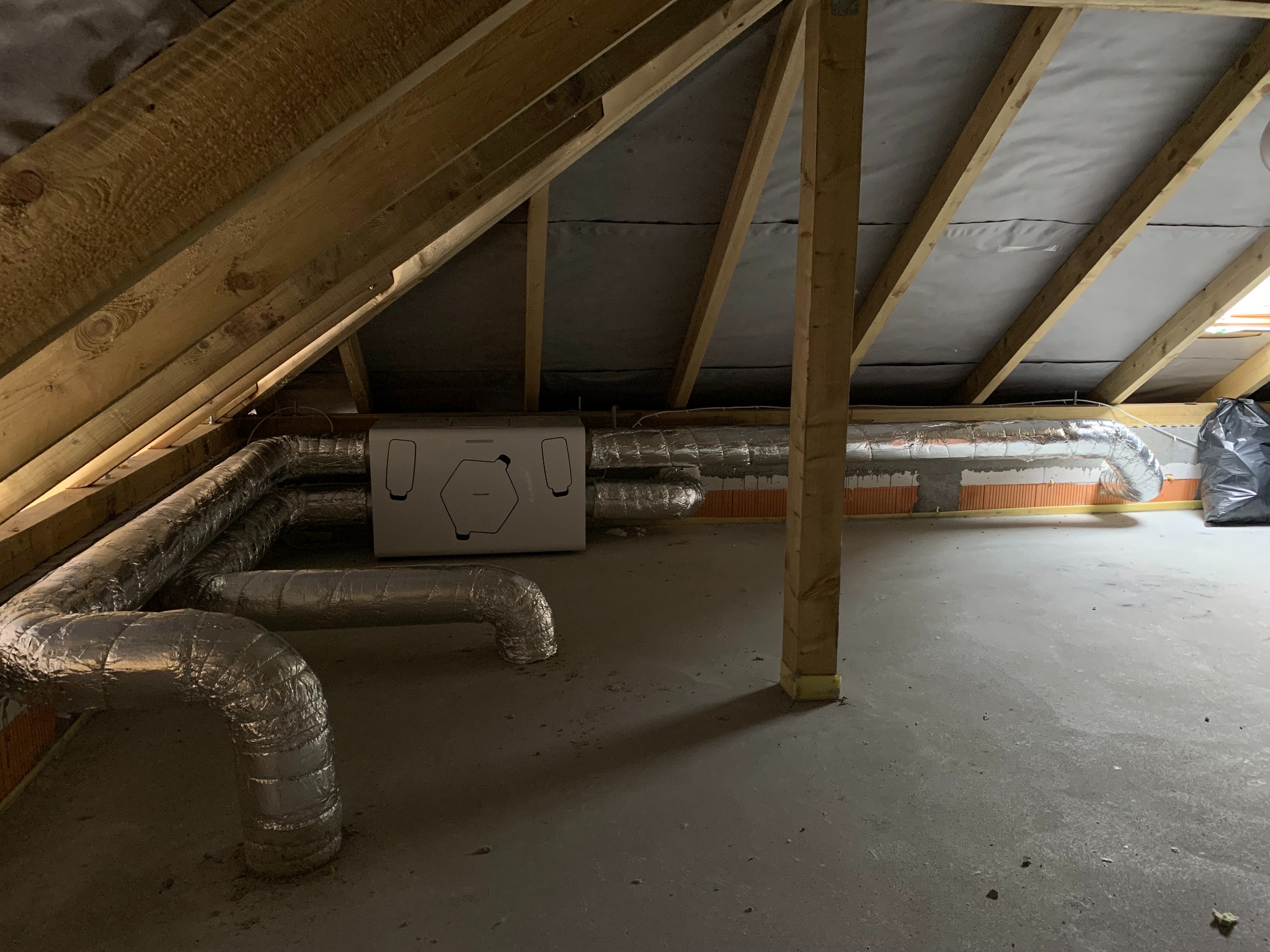 Instalacja wentylacyjna z centralą w surowym poddaszu, widoczne rury spiro owinięte izolacją, drewniana konstrukcja dachu i folia paroizolacyjna.