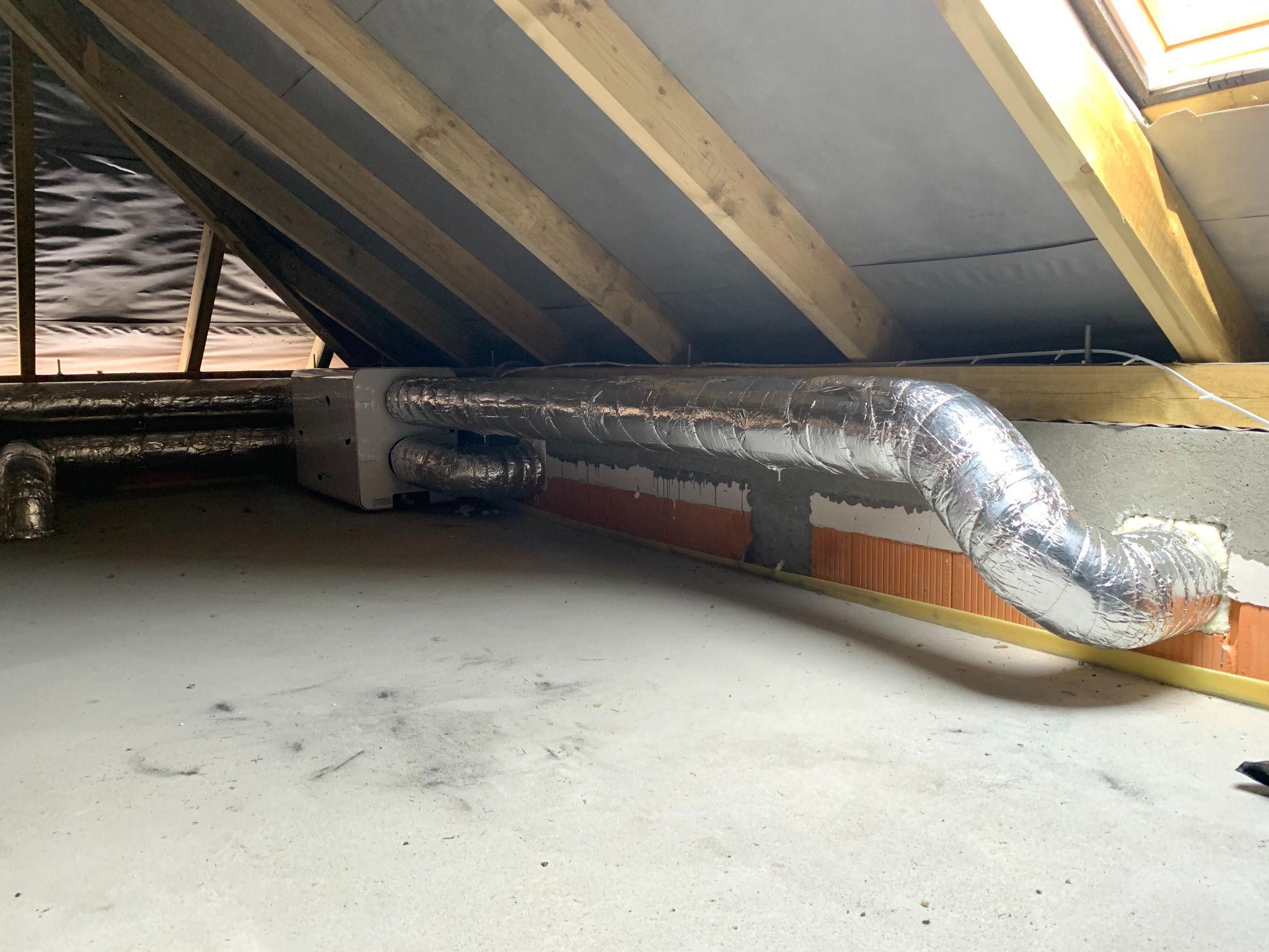 Instalacja wentylacyjna z aluminiowymi przewodami w przestrzeni poddasza, widoczne drewniane elementy konstrukcyjne dachu i fragment okna dachowego.