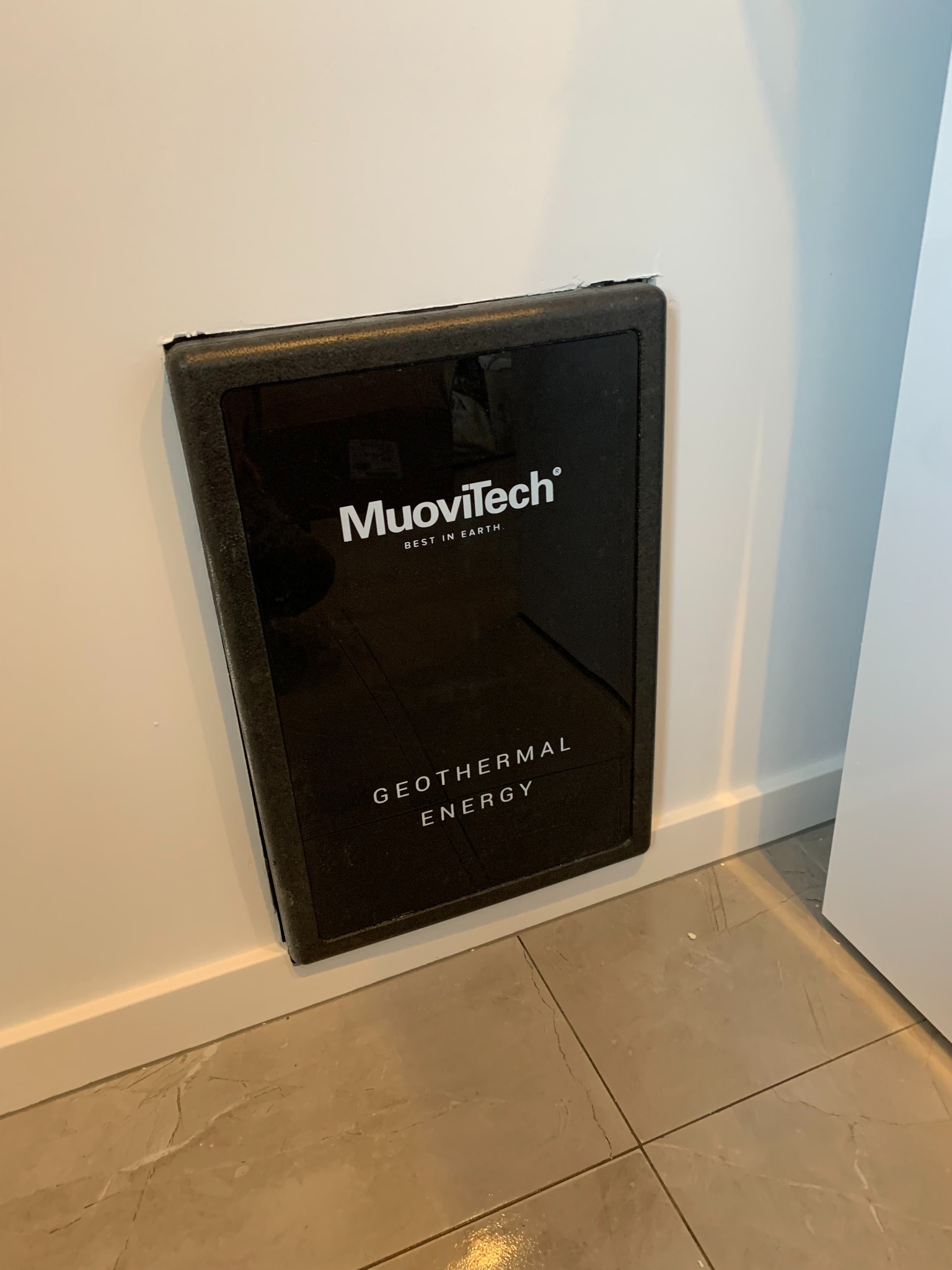 Czarna, prostokątna obudowa systemu geotermalnego MuoviTech, zamontowana przy podłodze na białej ścianie. Widoczna marmurowa podłoga i fragment drzwi.