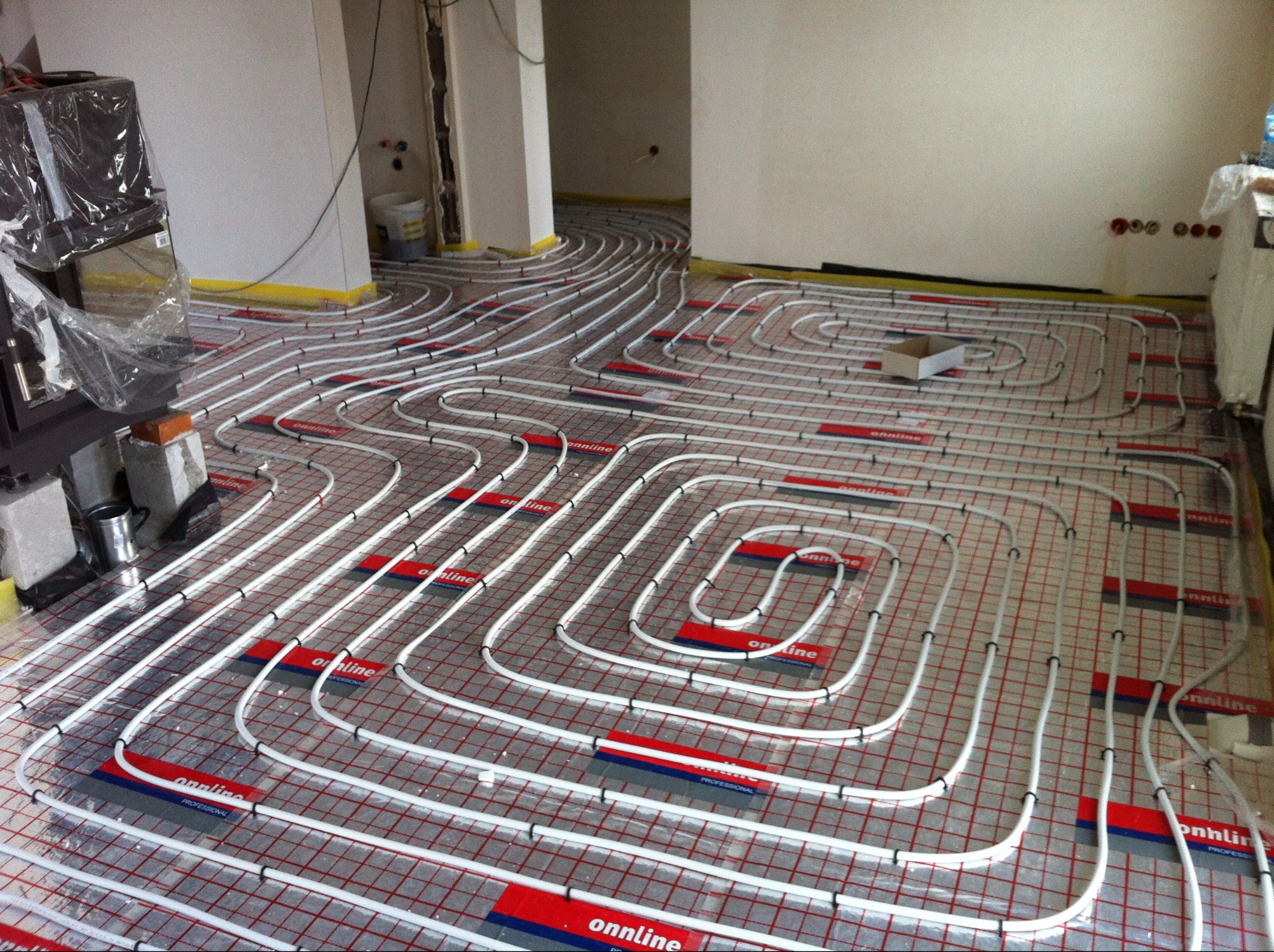 Białe rury ogrzewania podłogowego ułożone na czerwonej siatce w jasnym pomieszczeniu w trakcie instalacji. Widoczne elementy montażowe i przygotowane podłoże.