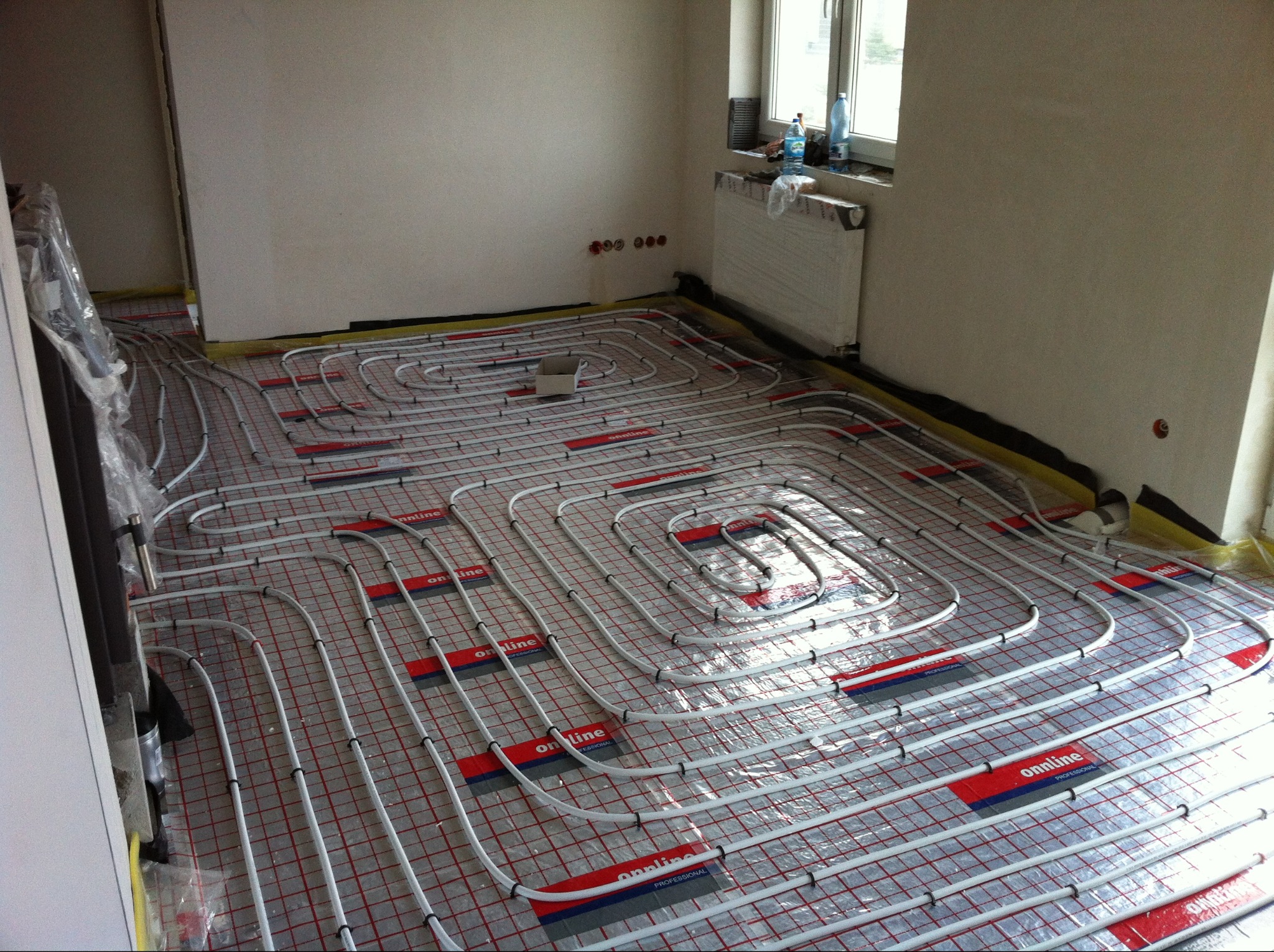 Ułożone rury ogrzewania podłogowego na folii z siatką, widoczne elementy mocujące. W tle okno z grzejnikiem i butelkami wody. Świeżo położona instalacja.
