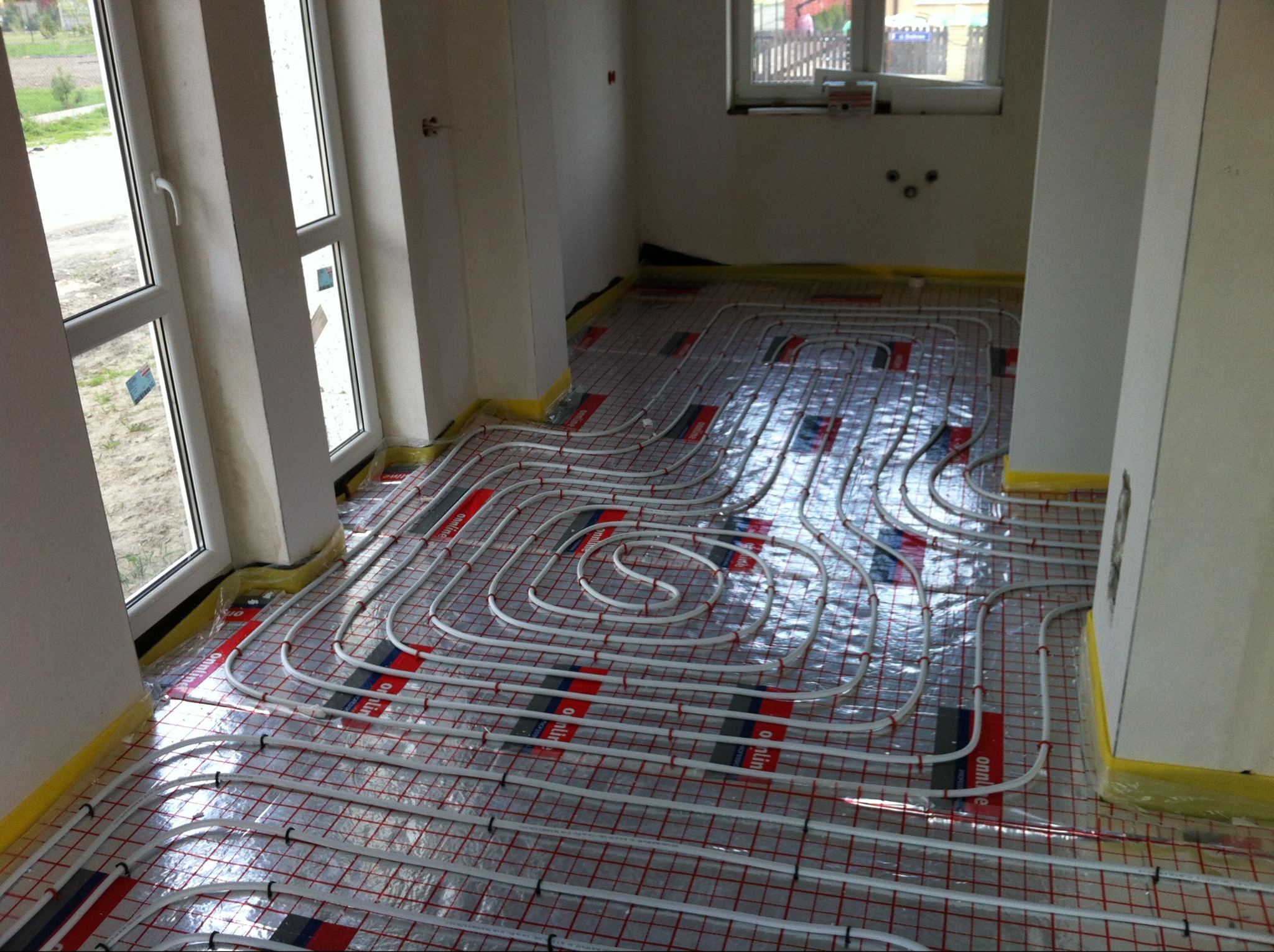 Białe rury ogrzewania podłogowego ułożone na folii z czerwoną kratką w surowym wnętrzu. Widoczne okna i fragmenty ścian. Żółta taśma zabezpiecza krawędzie.