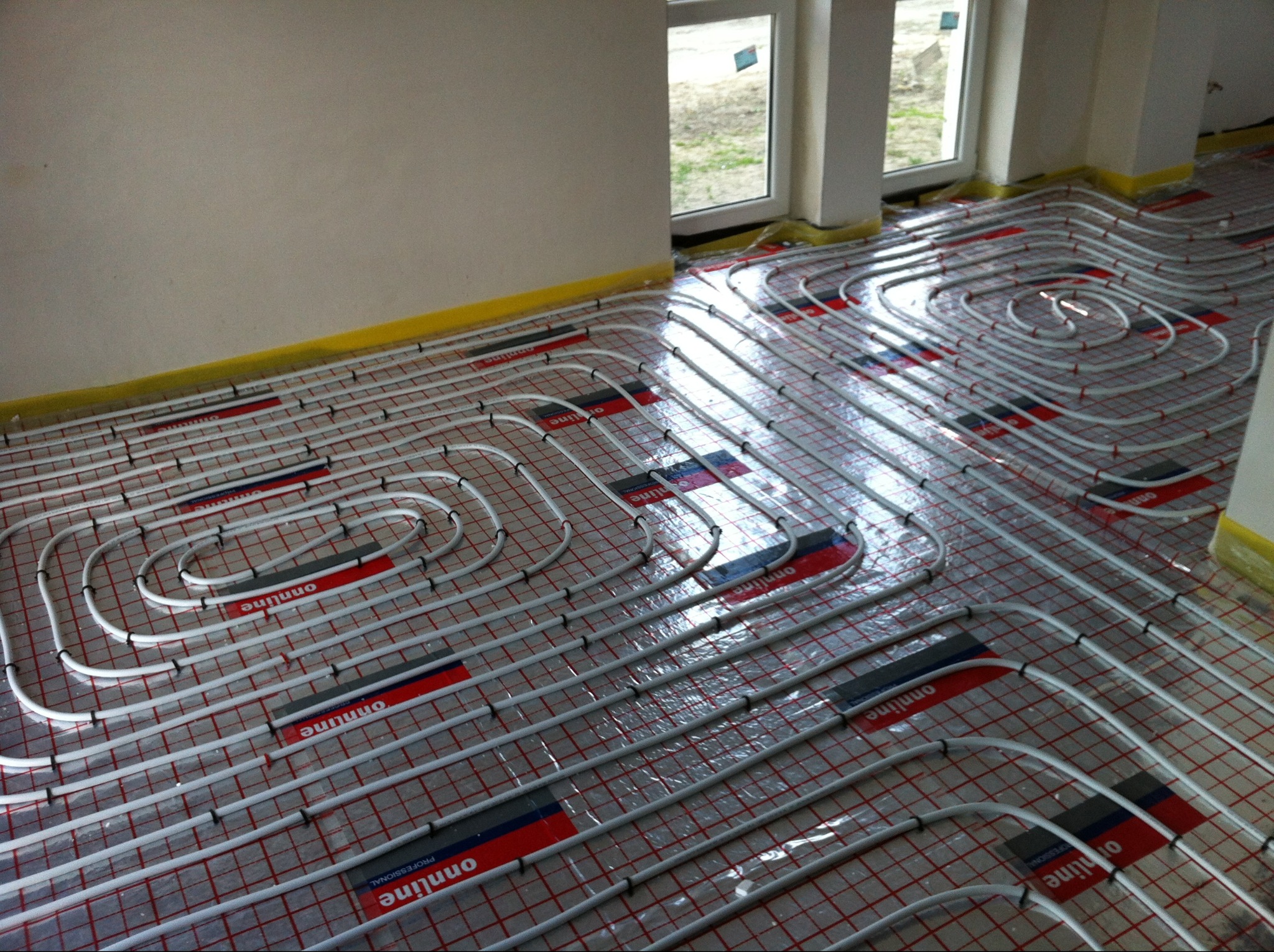 Białe rury ogrzewania podłogowego ułożone na czerwonej siatce, widoczne okno i fragment ściany. Żółta taśma zabezpieczająca przy ścianie. Ujęcie z góry, ukośne.