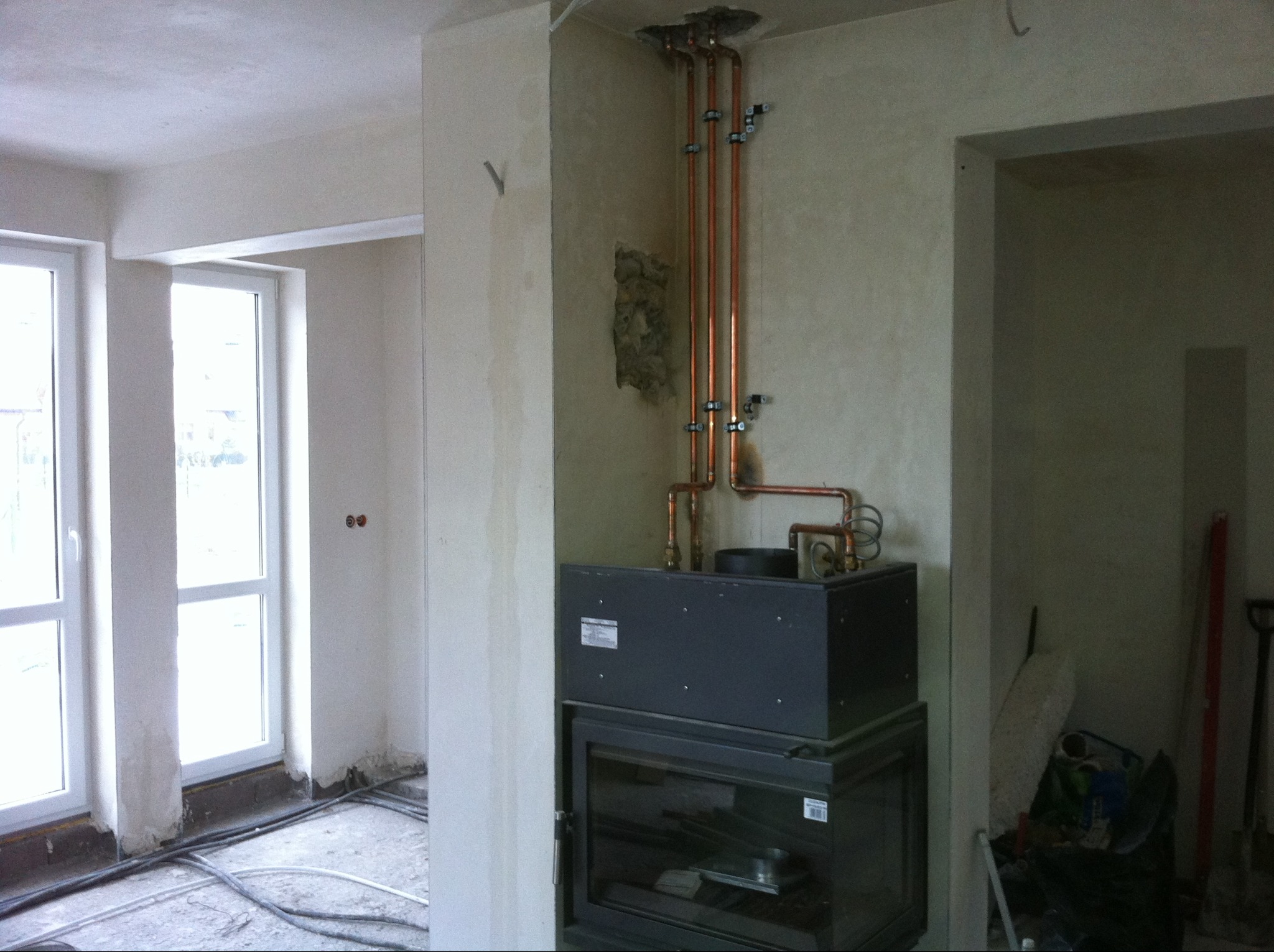 Montaż pieca z miedzianą instalacją w surowym wnętrzu. Widoczne rury, piec z szybą i fragmenty ścian w trakcie wykończenia. Prace instalacyjne w nowym budynku.