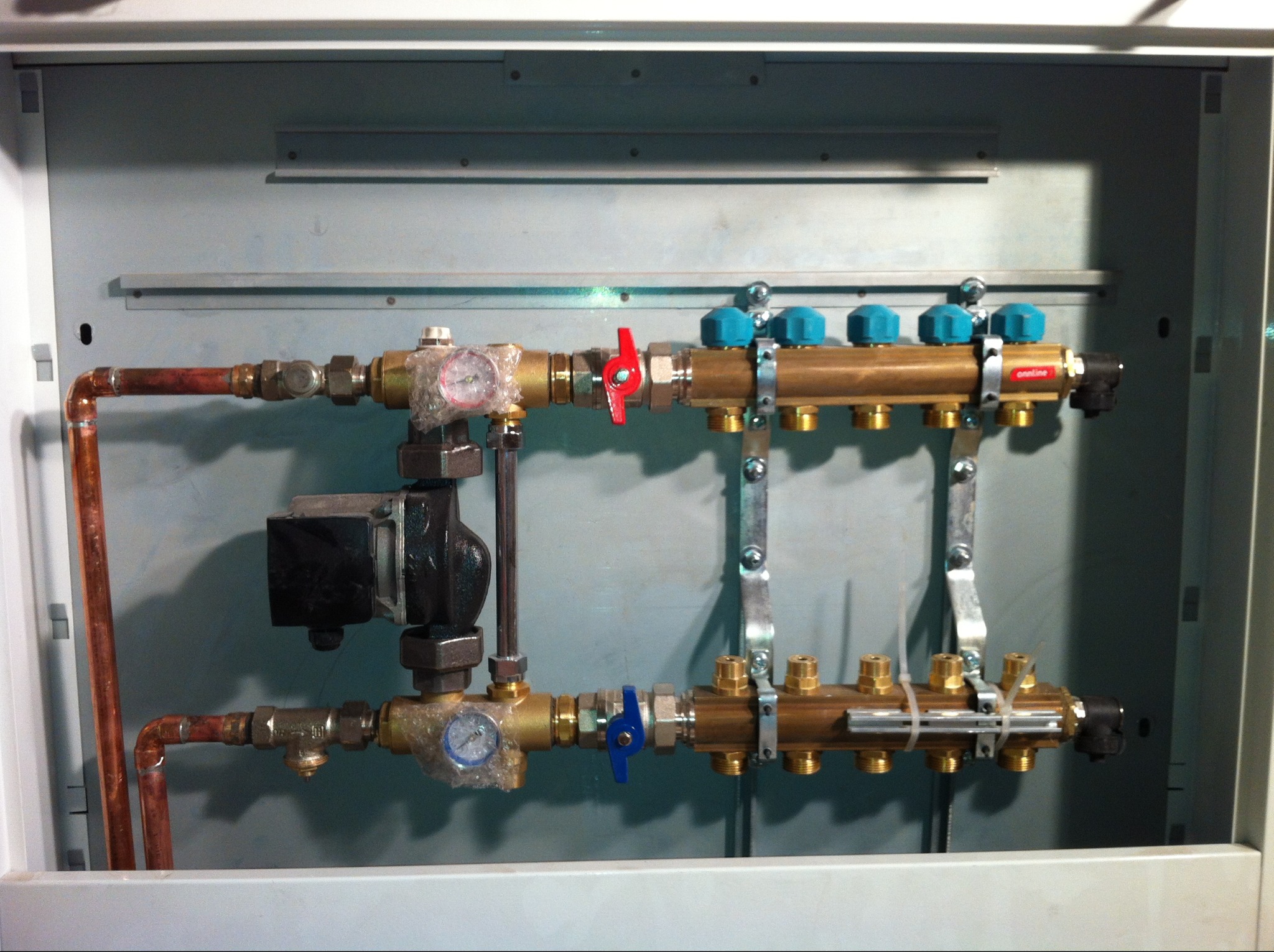 Rozdzielacz ogrzewania podłogowego w metalowej szafce, z miedzianymi rurami i zaworami z niebieskimi pokrętłami. Manometry zabezpieczone folią. Widoczne elementy instalacji.