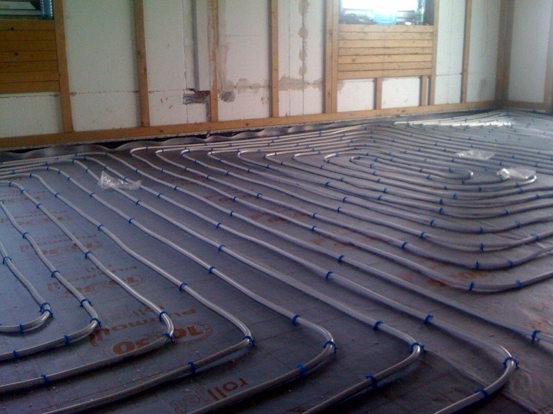 Ułożone rury ogrzewania podłogowego na izolacji, zabezpieczone niebieskimi uchwytami, widoczne fragmenty ścian w surowym stanie.
