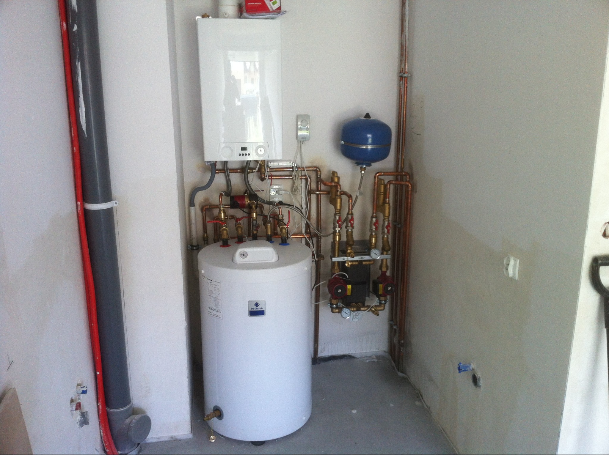 Biały piec gazowy De Dietrich z zbiornikiem i niebieskim naczyniem przeponowym, zamontowany w rogu pomieszczenia z widocznymi rurami miedzianymi i szarą rurą PCV.