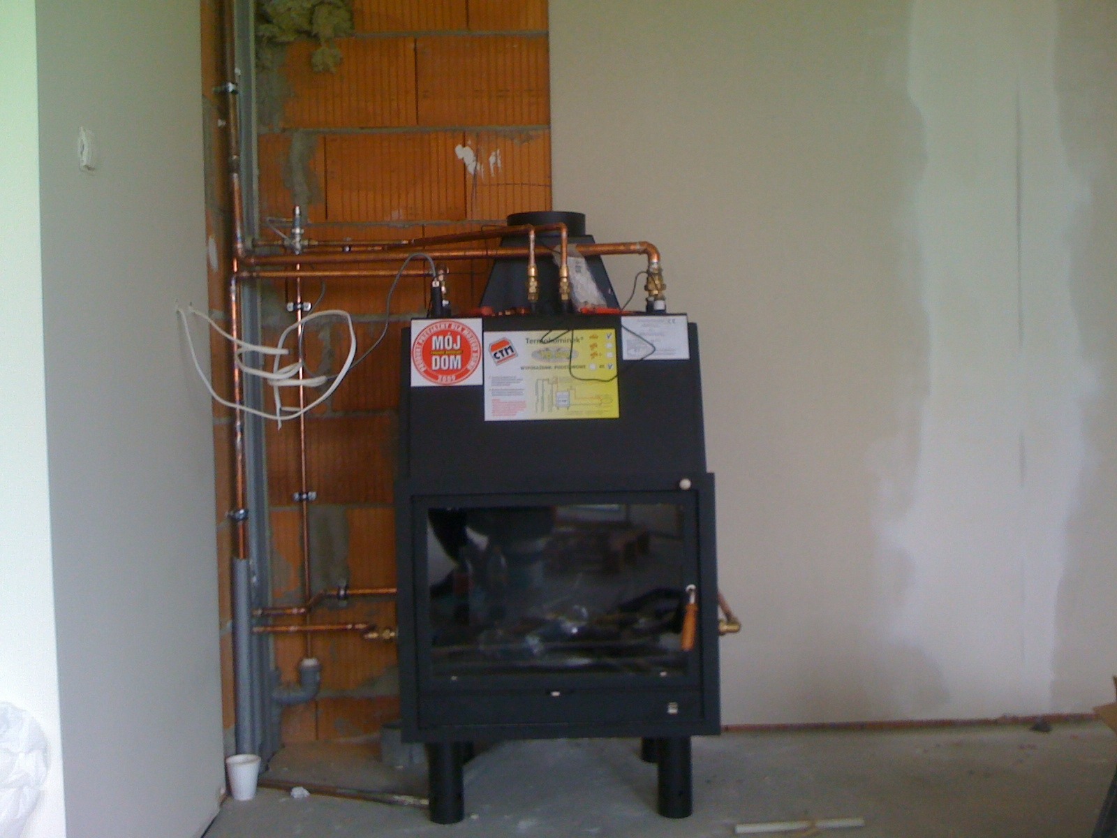 Czarny piec CO z instalacją miedzianą na ścianie z cegły. Widoczne rury, zawory i etykiety. Ściany w trakcie wykończenia.
