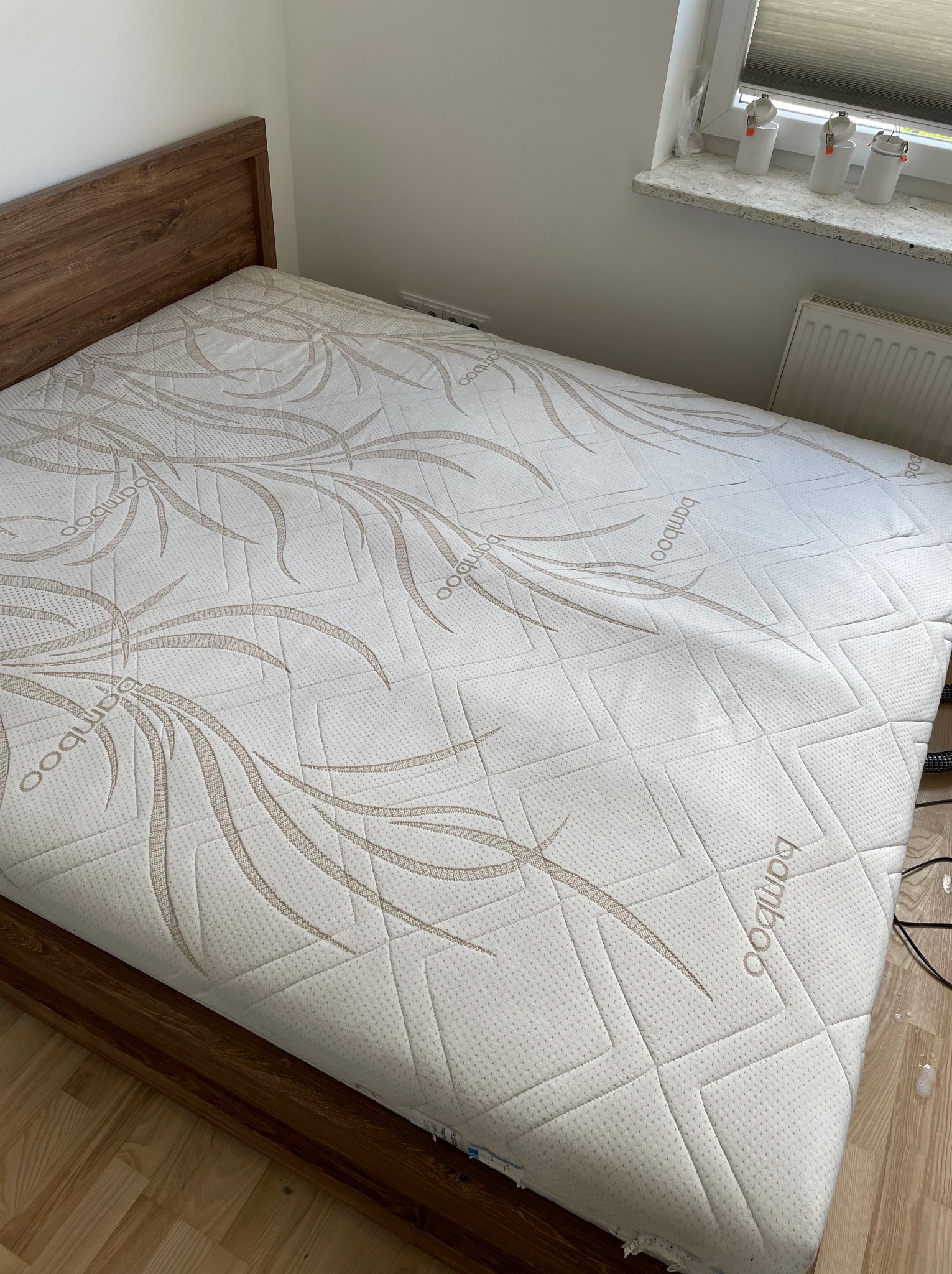 Materac na drewnianym łóżku z wzorem bambusa, w jasnym pokoju z oknem i grzejnikiem. Widoczne detale materaca i ramy łóżka.