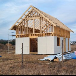 Piotr Krystkiewicz konstrukcje drewniane drewnianym2 - Surowy stan domu szkieletowego w trakcie budowy. Widoczne drewniane elementy konstrukcyjne i białe płyty elewacyjne. Na pierwszym planie trawa i ogrodzenie.