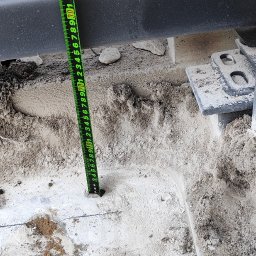 BRAMATIK Konrad Szewczuk - Pomiar głębokości otworu pod słupek bramy wjazdowej za pomocą miarki. Widoczne elementy konstrukcji bramy i przygotowany teren pod montaż.