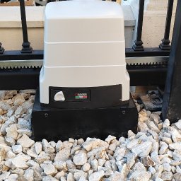 BRAMATIK Konrad Szewczuk - Zamontowany automat do bramy wjazdowej Roger na tle ozdobnych kamieni i fragmentu czarnej, metalowej bramy. Widoczny fragment samochodu za bramą.