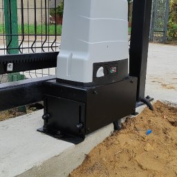 BRAMATIK Konrad Szewczuk - Zamontowany napęd bramy przesuwnej Roger na betonowej podstawie, widoczna listwa zębata i fragment ogrodzenia panelowego. Tło: dom i zieleń.