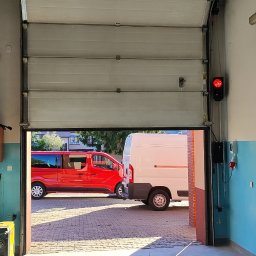 BRAMATIK - Otwarta segmentowa brama garażowa z sygnalizacją świetlną, widok z wnętrza garażu na podjazd z dwoma samochodami dostawczymi w słoneczny dzień.