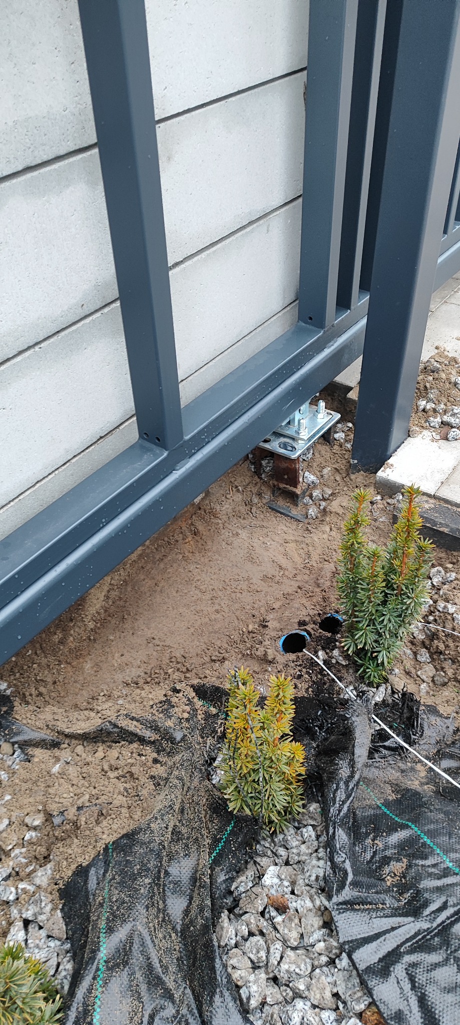 Szara brama przesuwna w trakcie montażu, widoczne elementy mocujące do podłoża, ziemia, kamienie i rośliny. Ujęcie z bliska, z dołu.