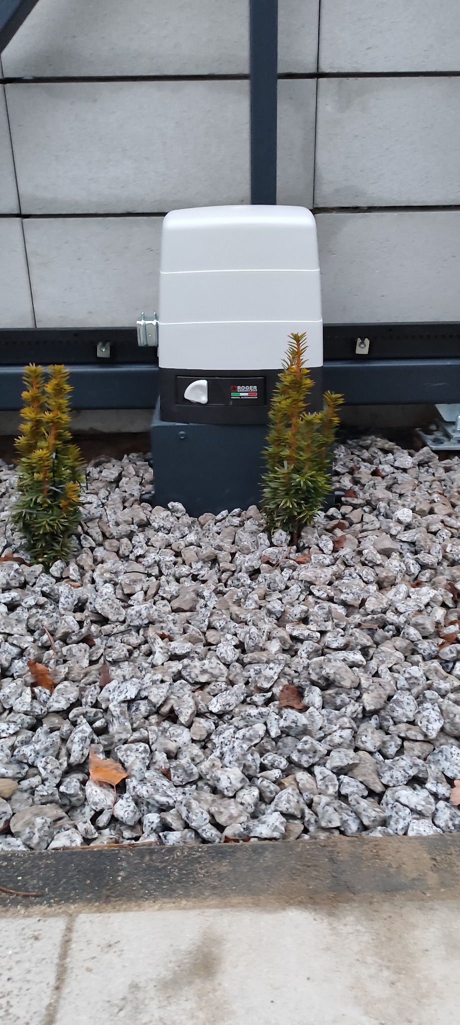 Zamontowany automat do bramy marki Roger, otoczony ozdobnymi kamieniami i dwoma małymi iglakami. Widoczna szyna bramy i fragment elewacji.