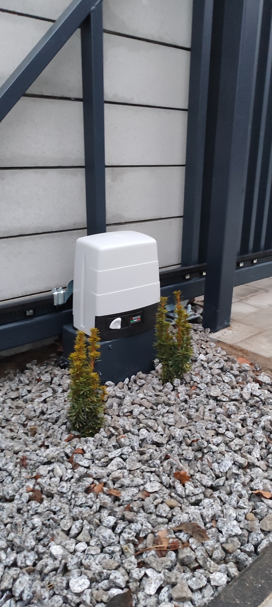 Napęd bramy przesuwnej z obudową w kolorze białym, zamontowany przy szarej bramie, otoczony ozdobnym kamieniem i dwoma małymi iglakami.