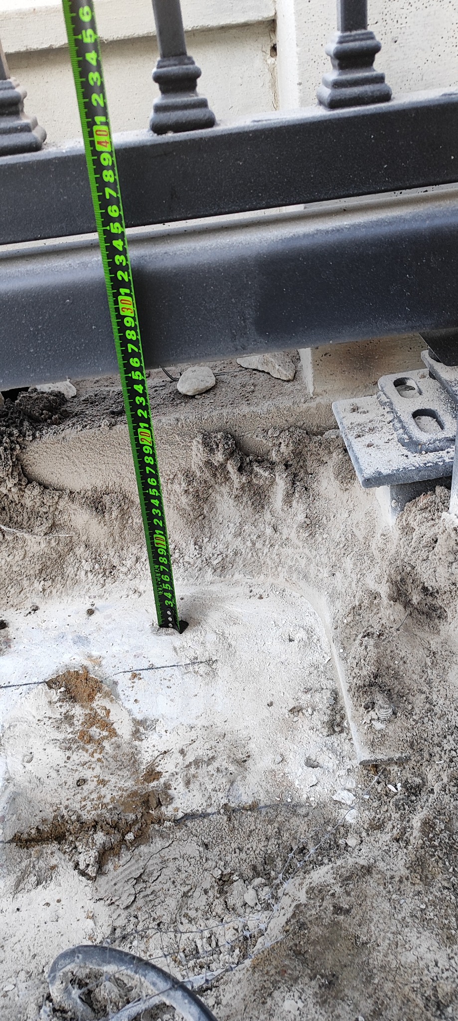 Pomiar głębokości otworu pod słupek bramy wjazdowej za pomocą miarki. Widoczne elementy konstrukcji bramy i przygotowany teren pod montaż.