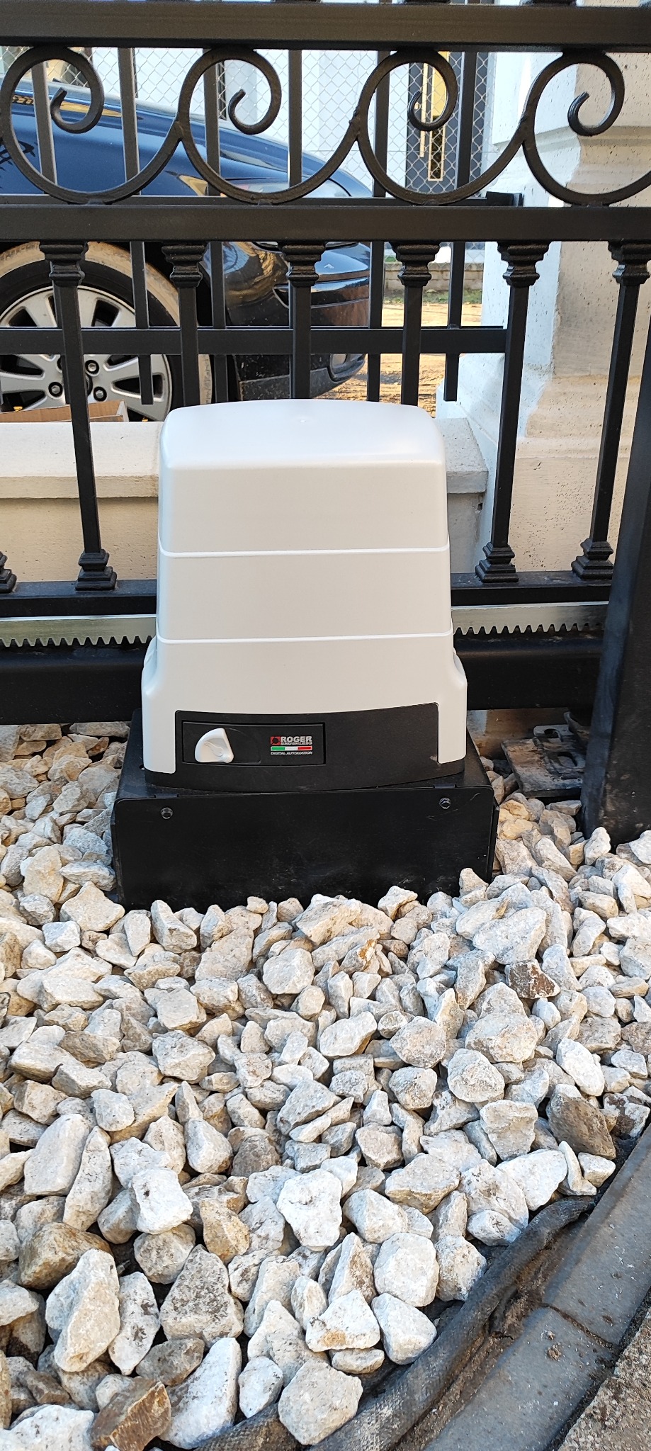 Zamontowany automat do bramy wjazdowej Roger na tle ozdobnych kamieni i fragmentu czarnej, metalowej bramy. Widoczny fragment samochodu za bramą.