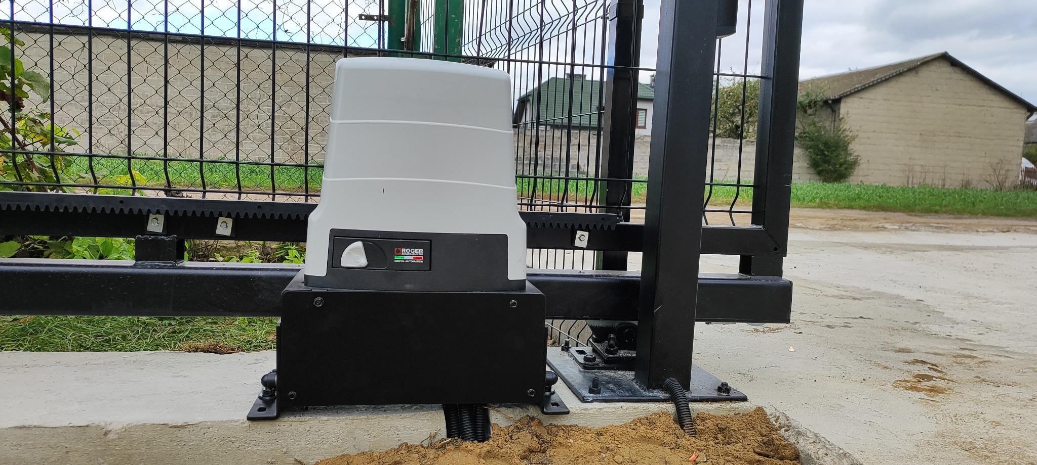 Zamontowany napęd bramy przesuwnej Roger na betonowej podstawie, widoczna zębatka i fragment ogrodzenia panelowego. Tło stanowi budynek gospodarczy i pochmurne niebo.