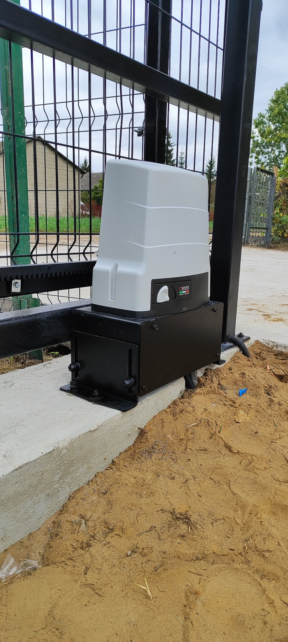 Zamontowany napęd bramy przesuwnej Roger na betonowej podstawie, widoczna listwa zębata i fragment ogrodzenia panelowego. Tło: dom i zieleń.