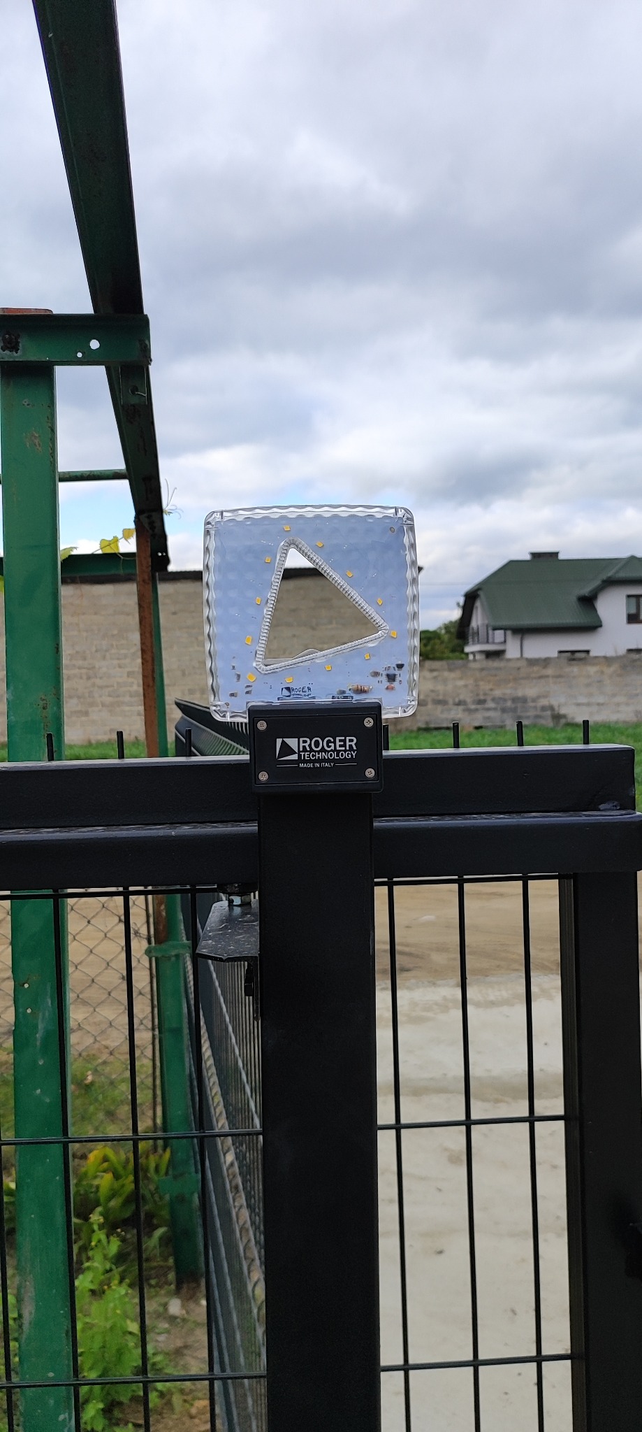 Lampa ostrzegawcza LED Roger Technology zamontowana na czarnej bramie wjazdowej, z widocznym domem w tle i pochmurnym niebem.
