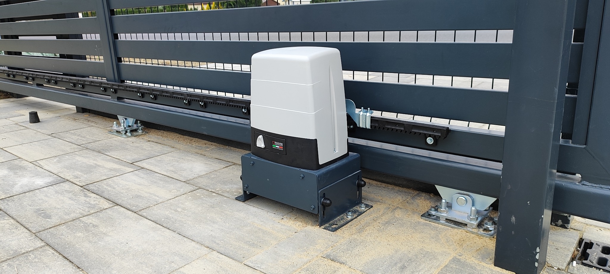 Szara brama przesuwna z zamontowanym automatem Roger na kostce brukowej. Widoczne elementy mocujące i listwa zębata. Całość w neutralnej kolorystyce.