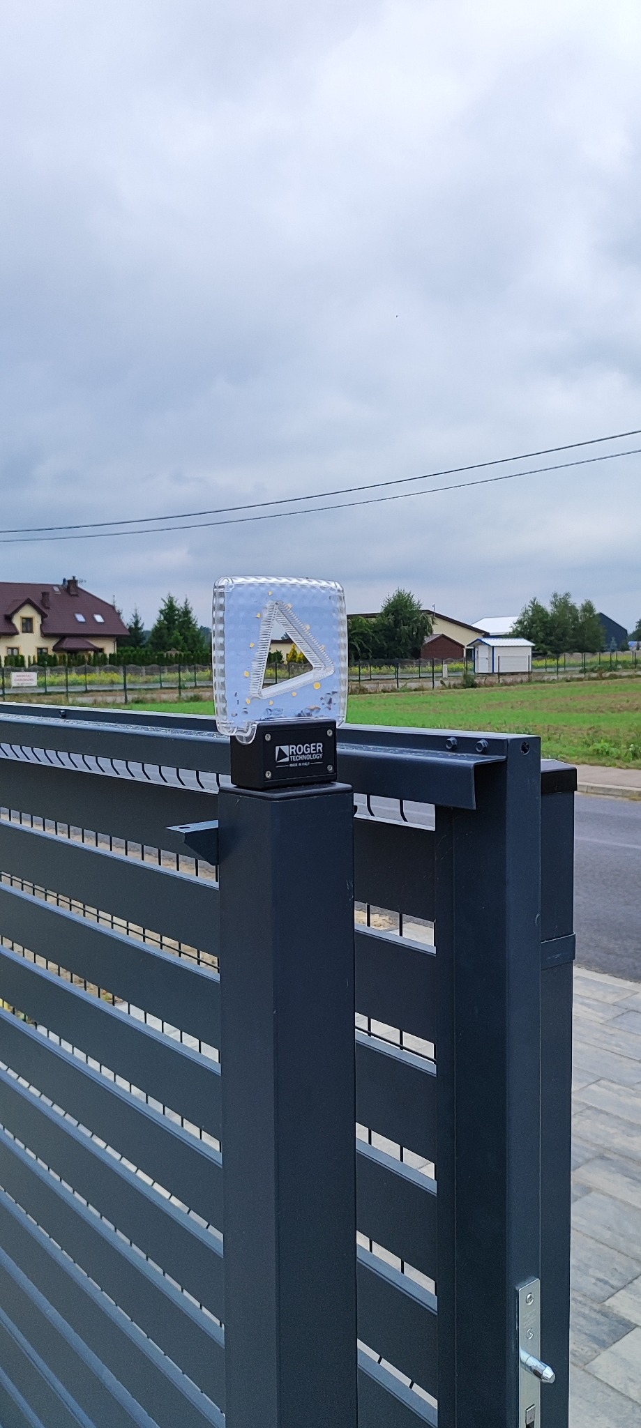Szara brama wjazdowa z lampą LED marki Roger Technology. Widoczne domy w tle i pochmurne niebo. Brama panelowa z poziomymi szczeblami.