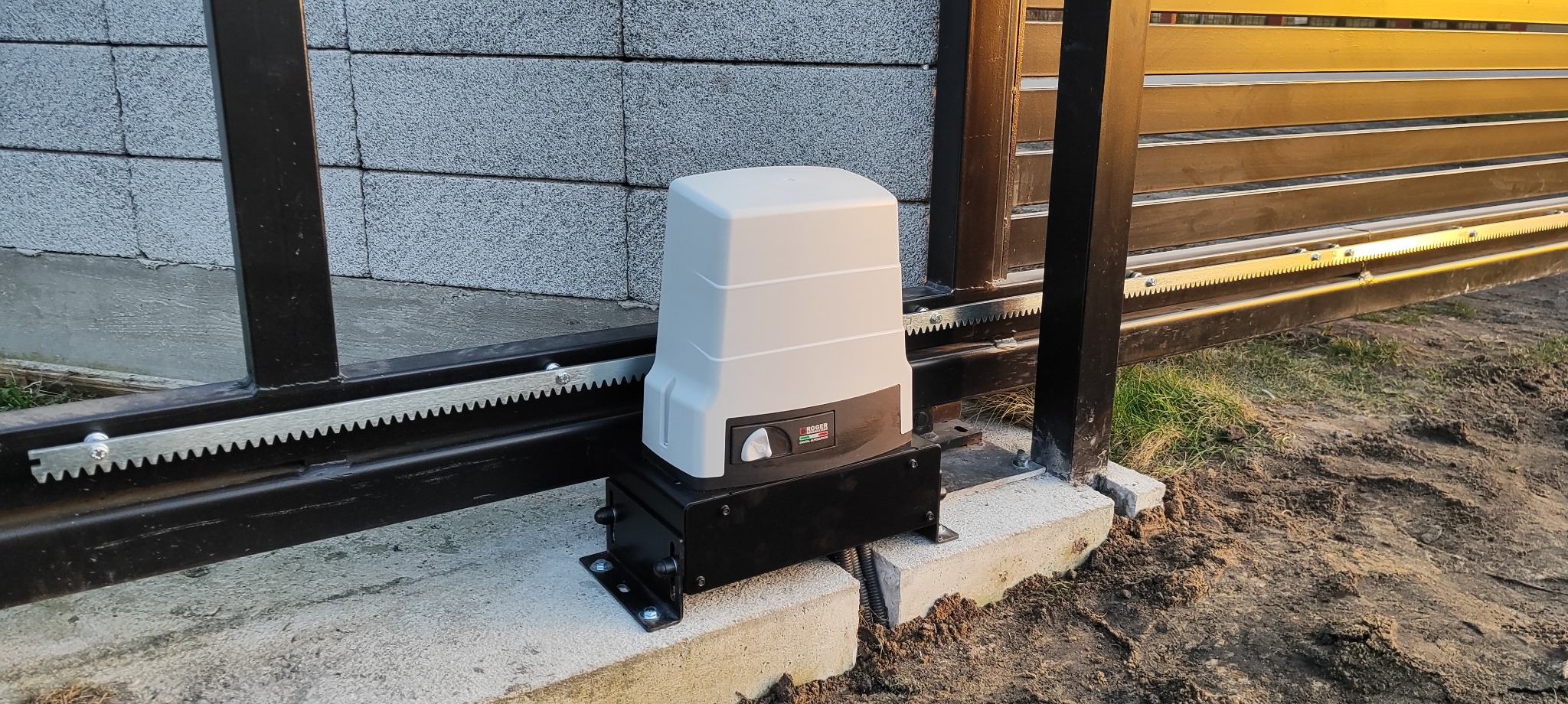 Zamontowany napęd bramy przesuwnej Roger na betonowym fundamencie, widoczna listwa zębata i fragment bramy wjazdowej. Ujęcie z poziomu gruntu.