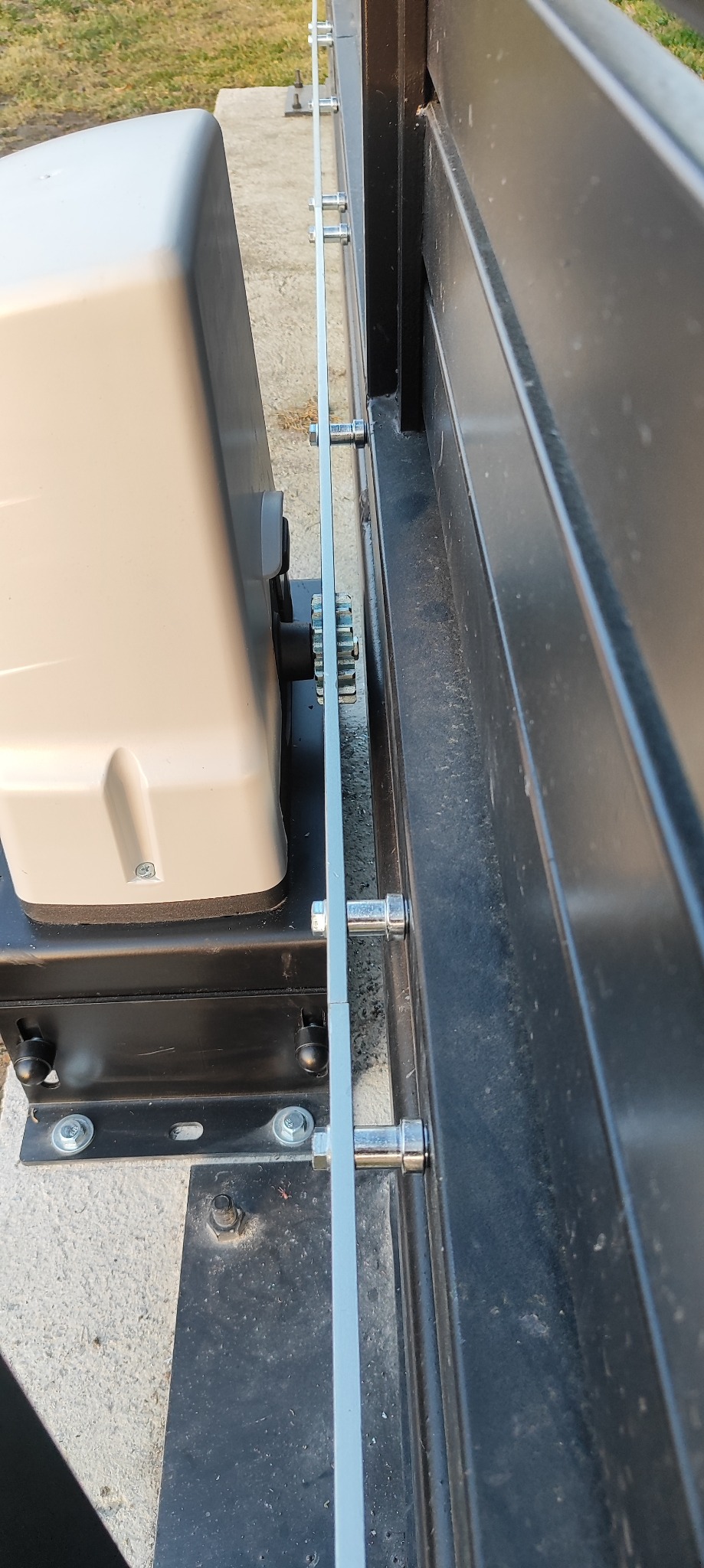 Szczegółowe ujęcie mechanizmu automatycznego otwierania bramy wjazdowej, z widocznym silnikiem, listwą zębatą i elementami mocującymi. Widok z boku, na zewnątrz.