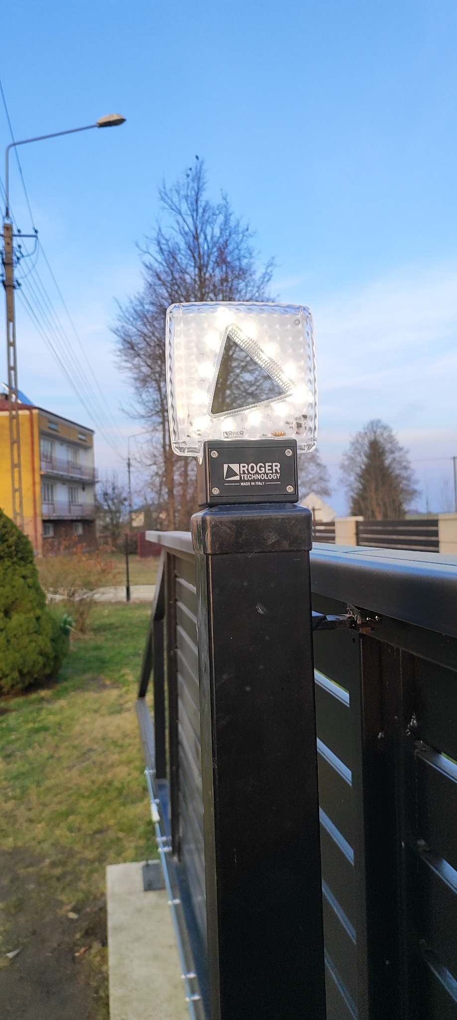 Lampa ostrzegawcza LED marki Roger Technology zamontowana na słupku czarnej bramy wjazdowej. Widoczne budynki w tle i błękitne niebo.