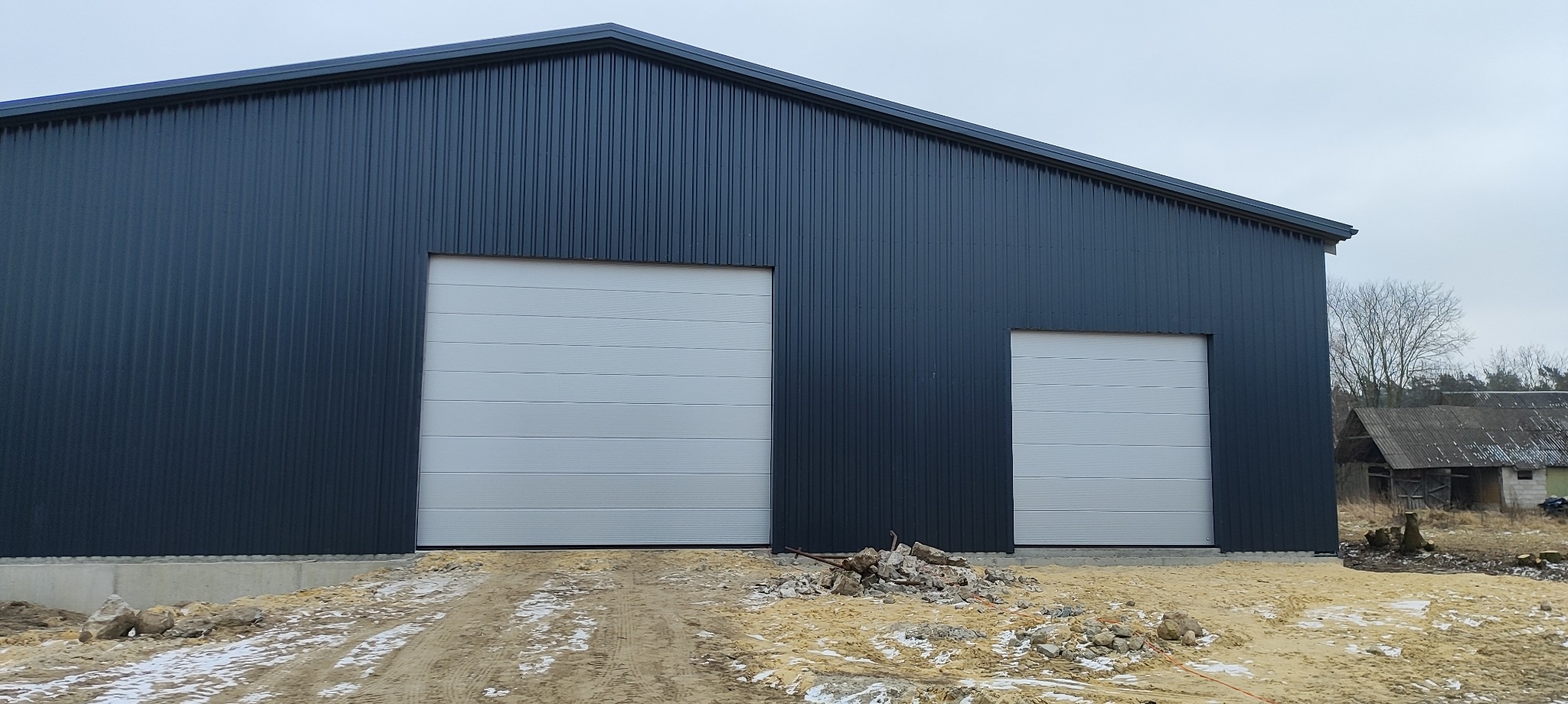 Nowoczesny budynek przemysłowy z dwiema segmentowymi bramami w kolorze białym. Elewacja z blachy trapezowej w odcieniach szarości. Teren wokół w trakcie prac wykończeniowych.